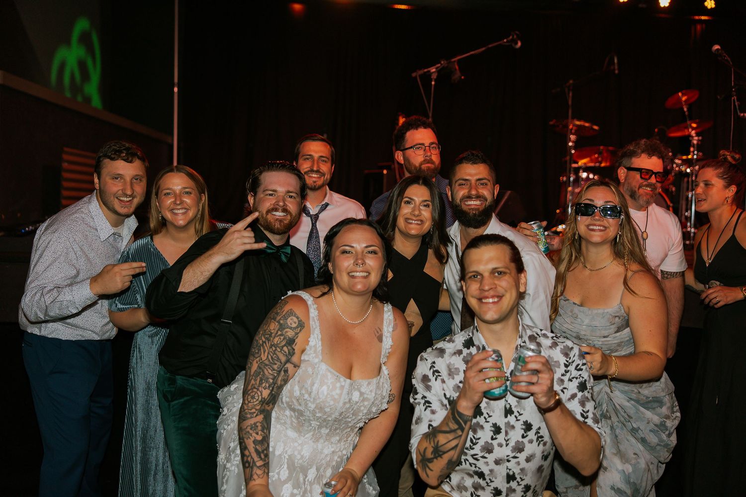 Group of wedding guests pose together smiling at reception venue with stage backdrop.