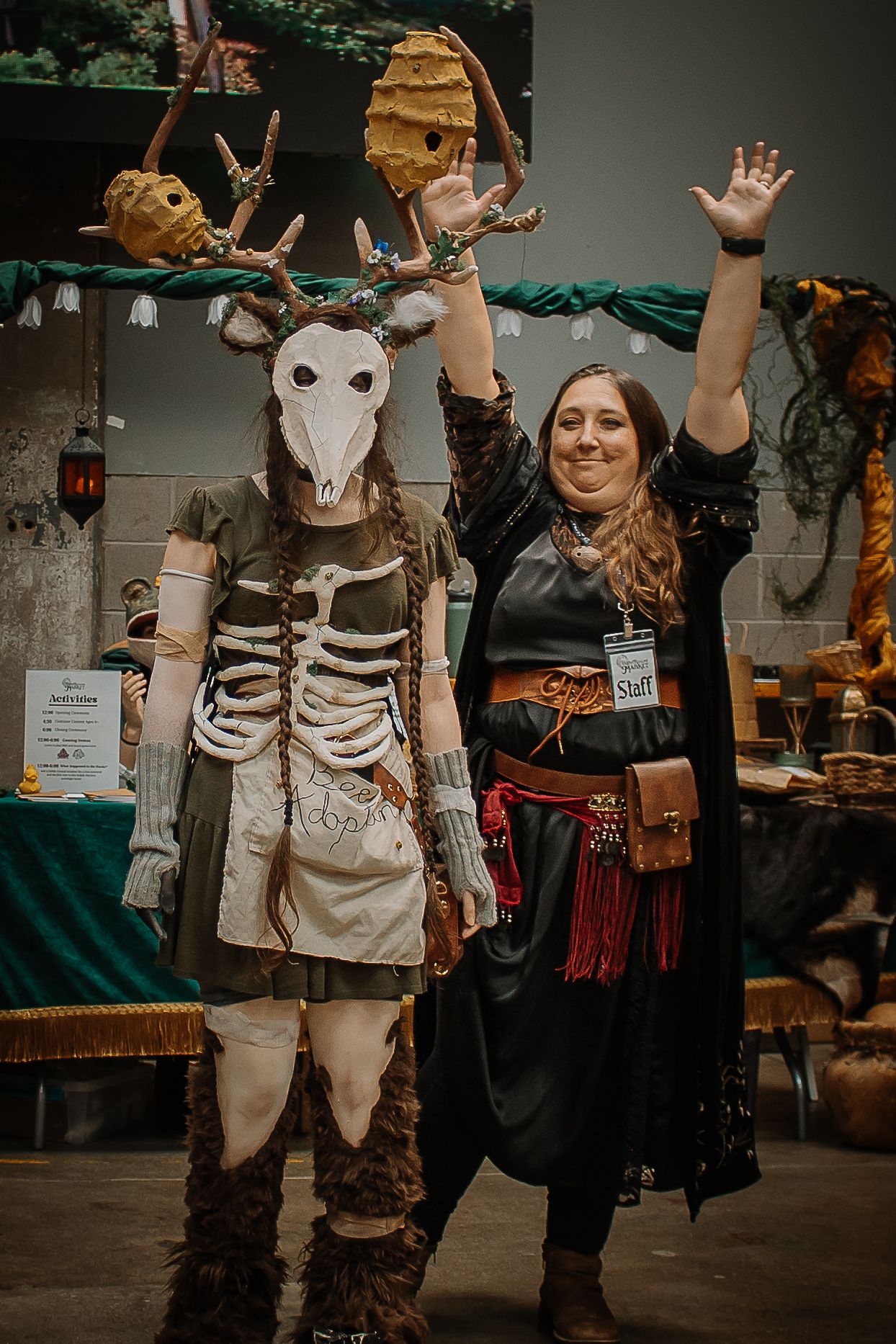 Two cosplayers at a convention, one in an elaborate deer skull and skeleton costume, the other in pirate attire with hands raised.