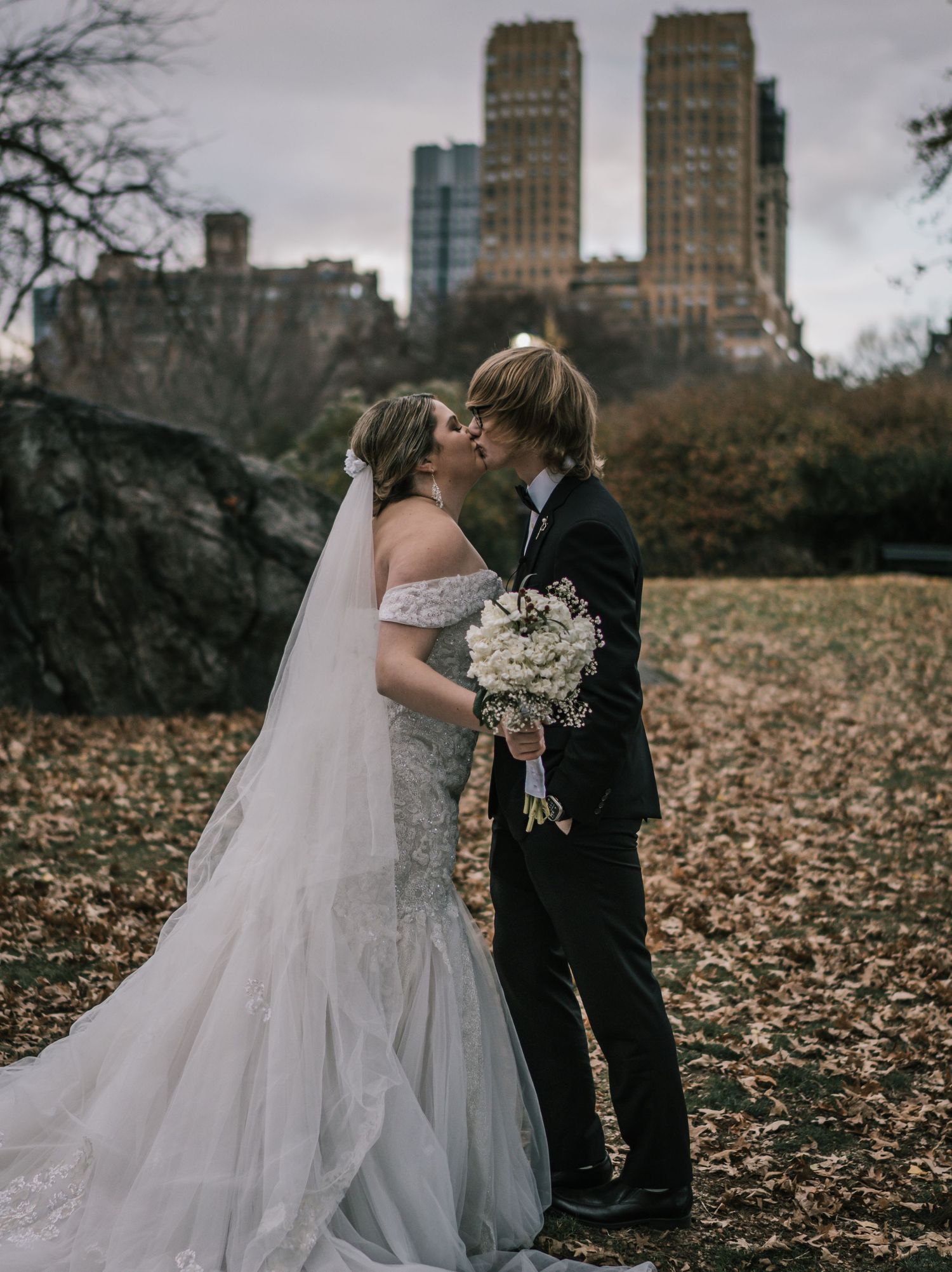 Bride and groom kiss holding white floral bouquet with NYC skyscrapers visible behind them in Central Park.