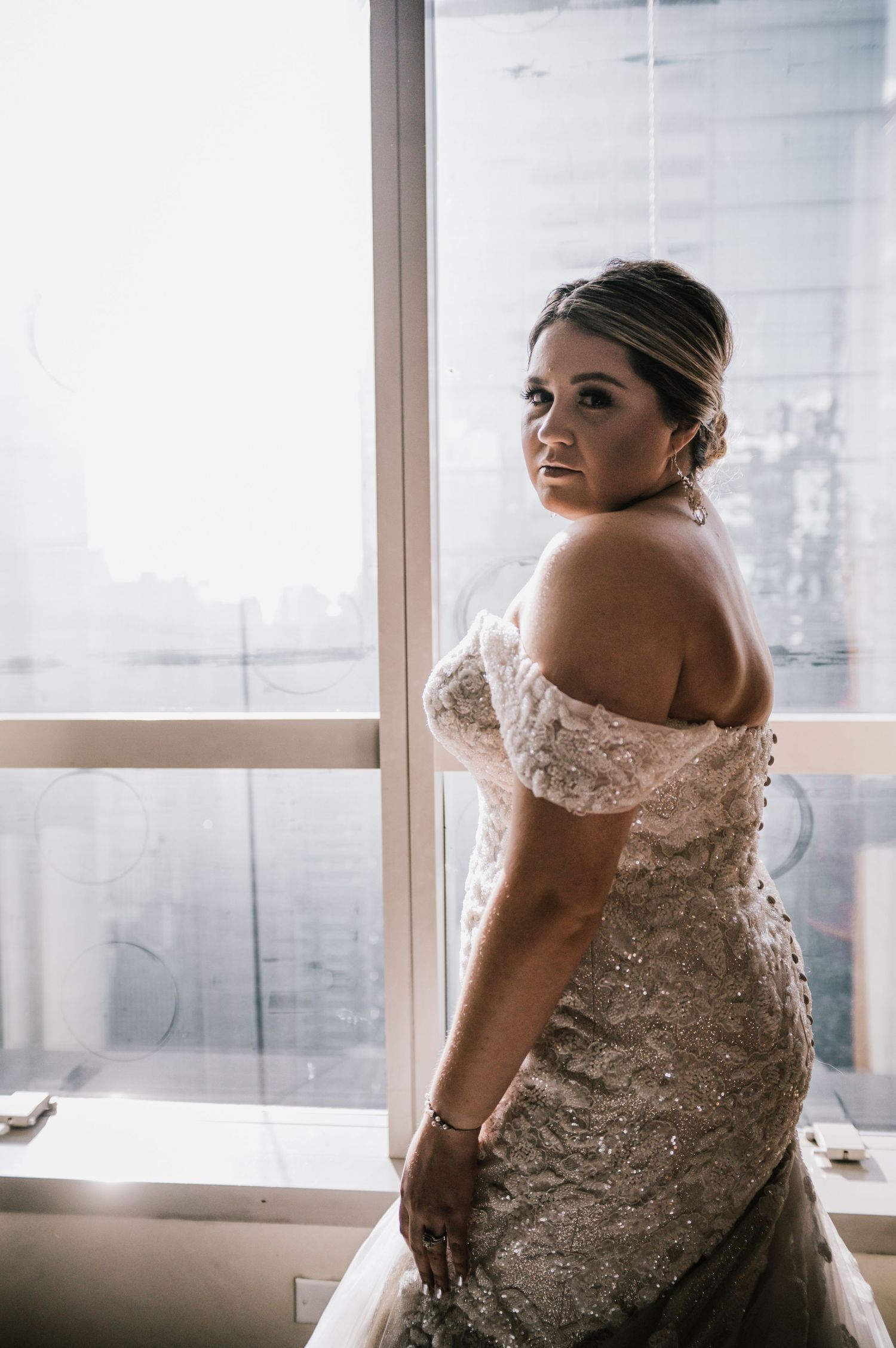 Bride in off-shoulder beaded mermaid wedding gown poses by window with city skyline backdrop.