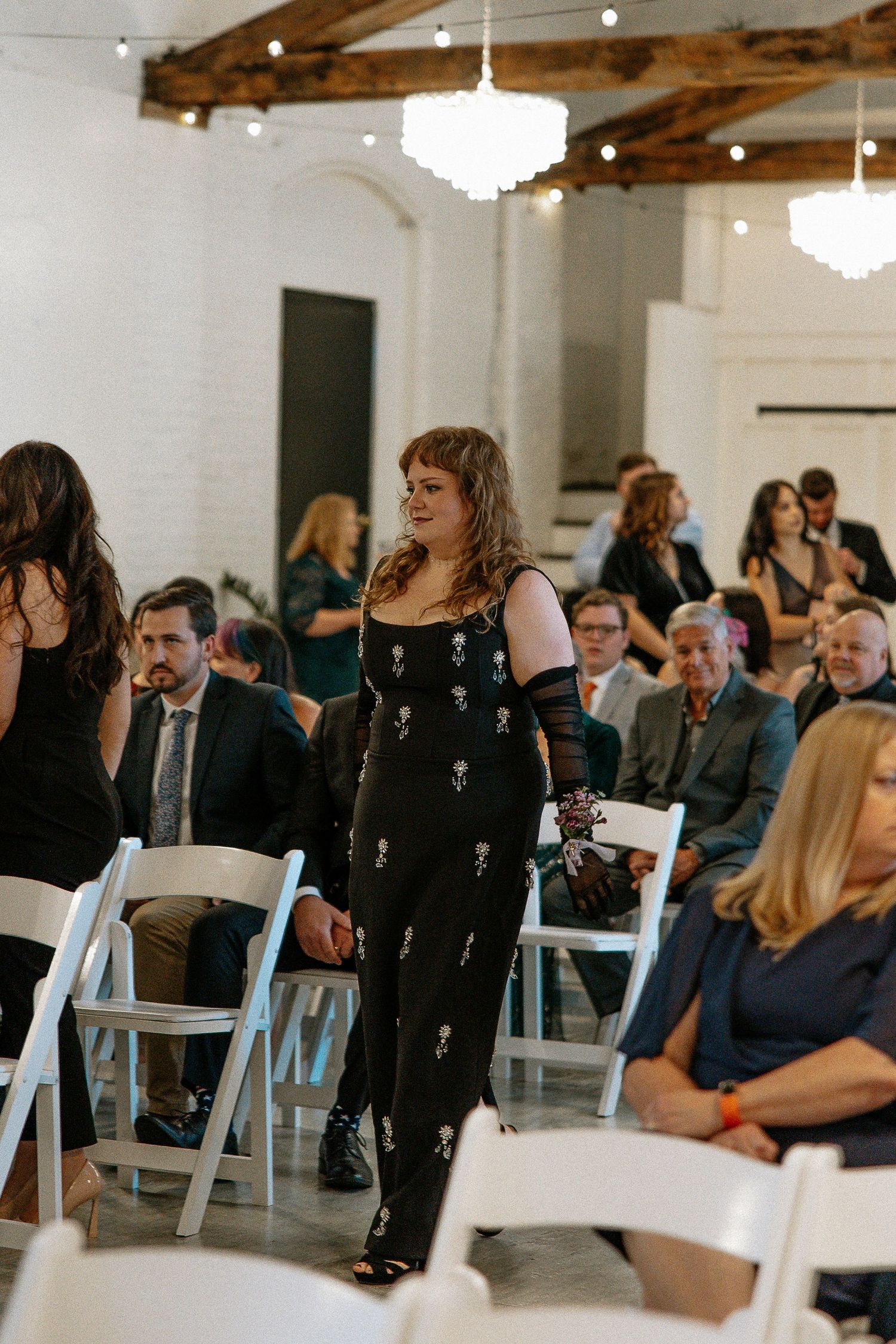 Wedding guest in black floral dress walking down aisle past seated guests in bright industrial venue.