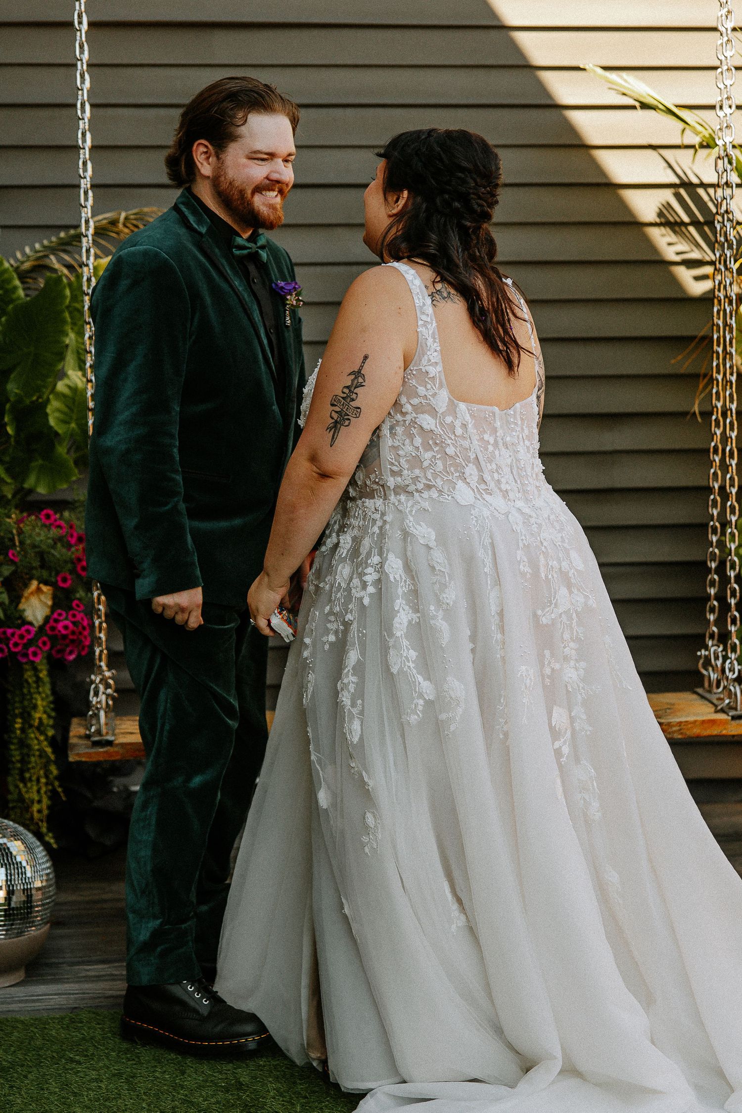 Bride and groom hold hands smiling at each other outdoors, she wears lace gown, he wears green velvet suit.