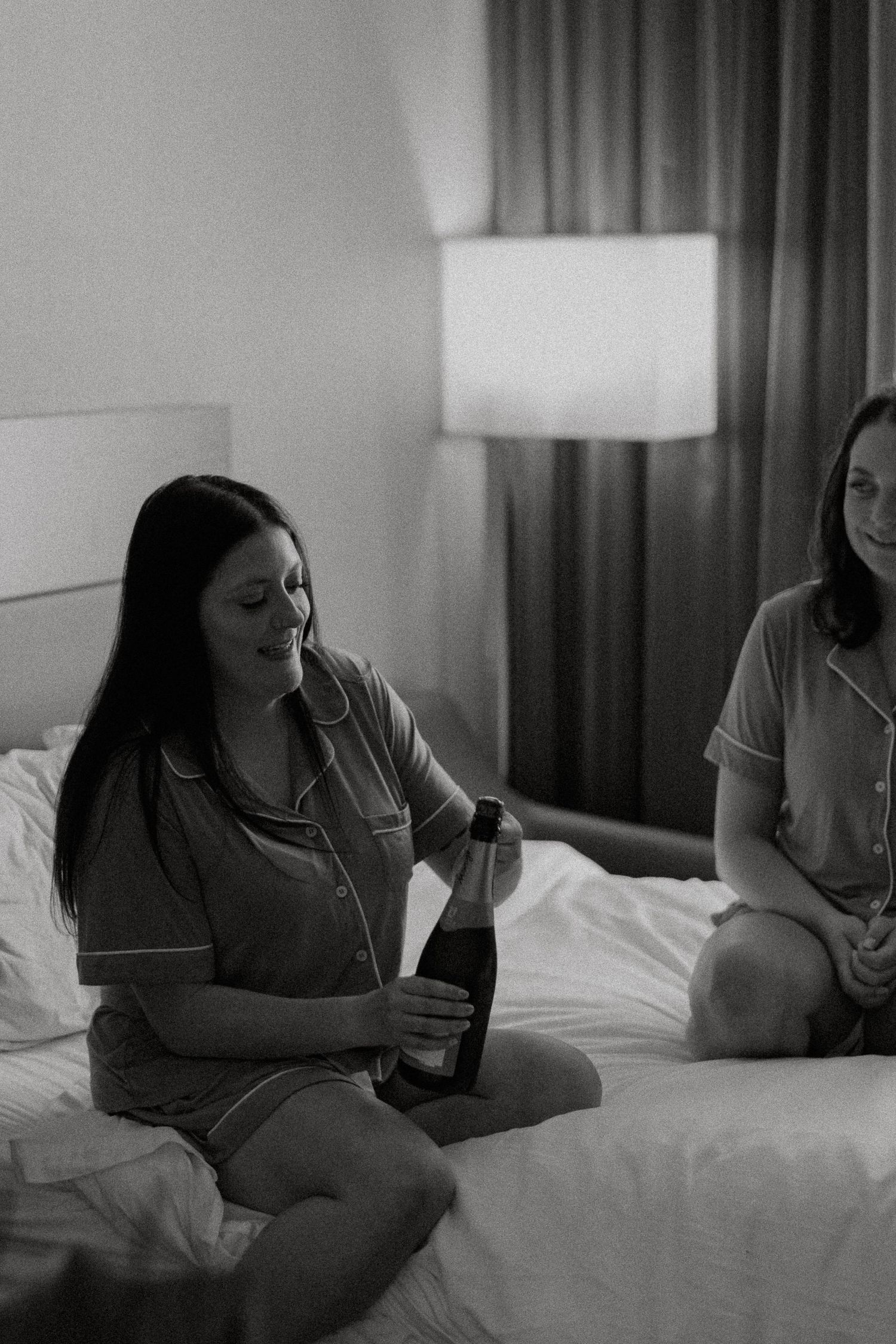 Black and white photo of woman in pajamas opening champagne bottle on hotel bed before wedding.