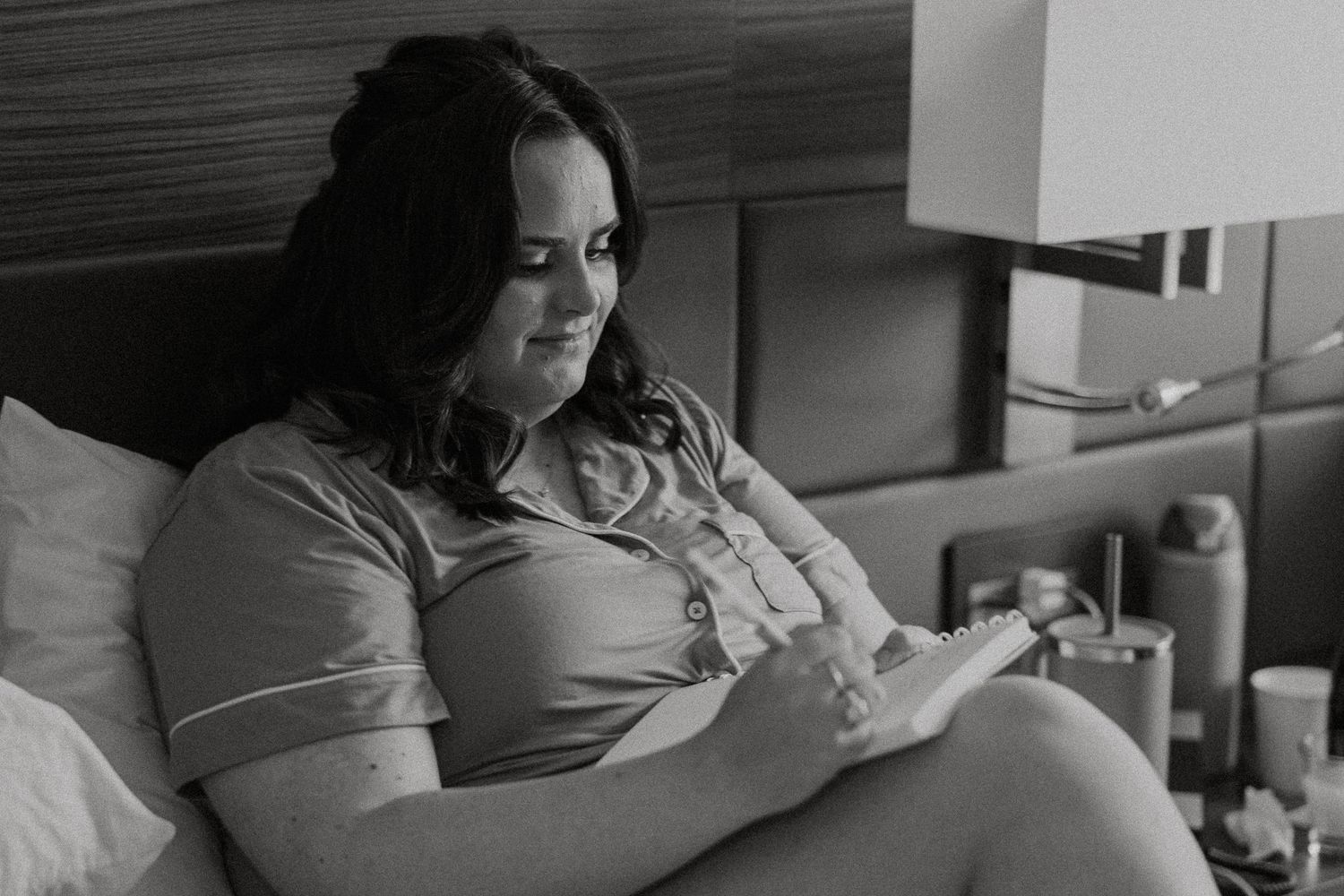 Black and white photo of a bride in green pajamas reading vows on a hotel bed before her wedding.