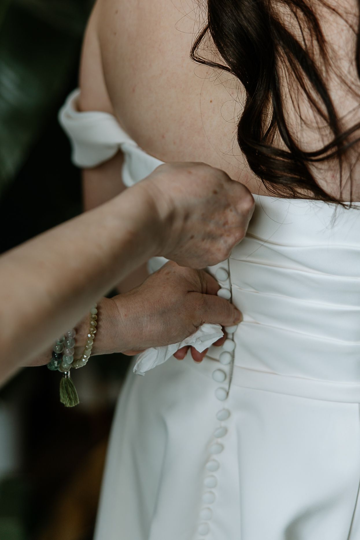 Close-up of mother fastening pearl buttons on back of bride's white wedding dress.