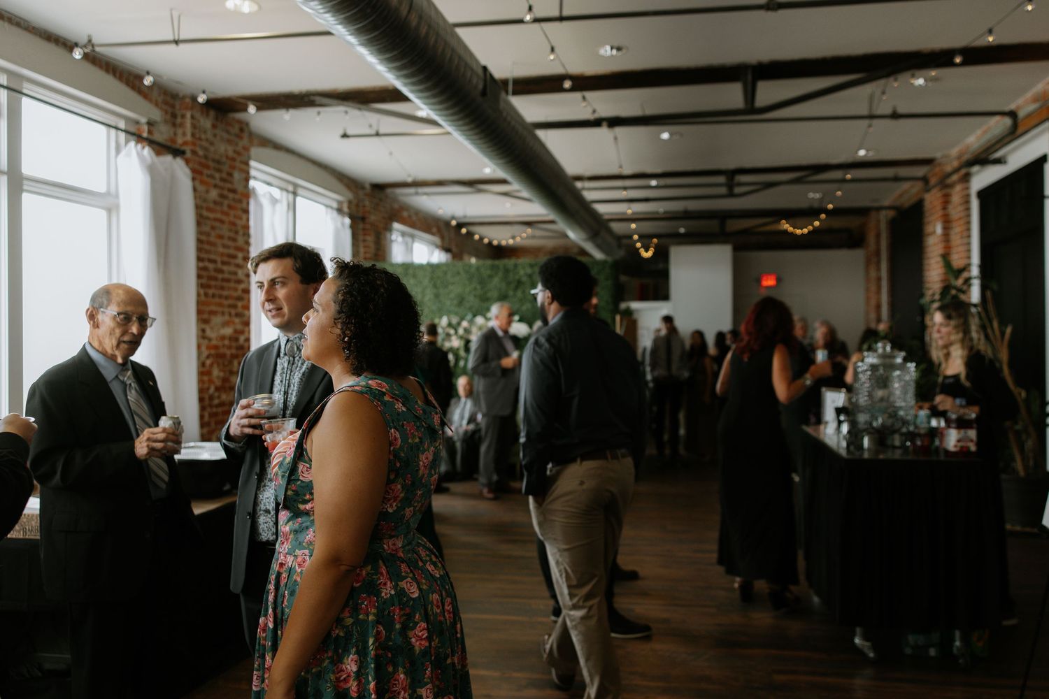Wedding cocktail hour guests mingling in a rustic brick venue with string lights.