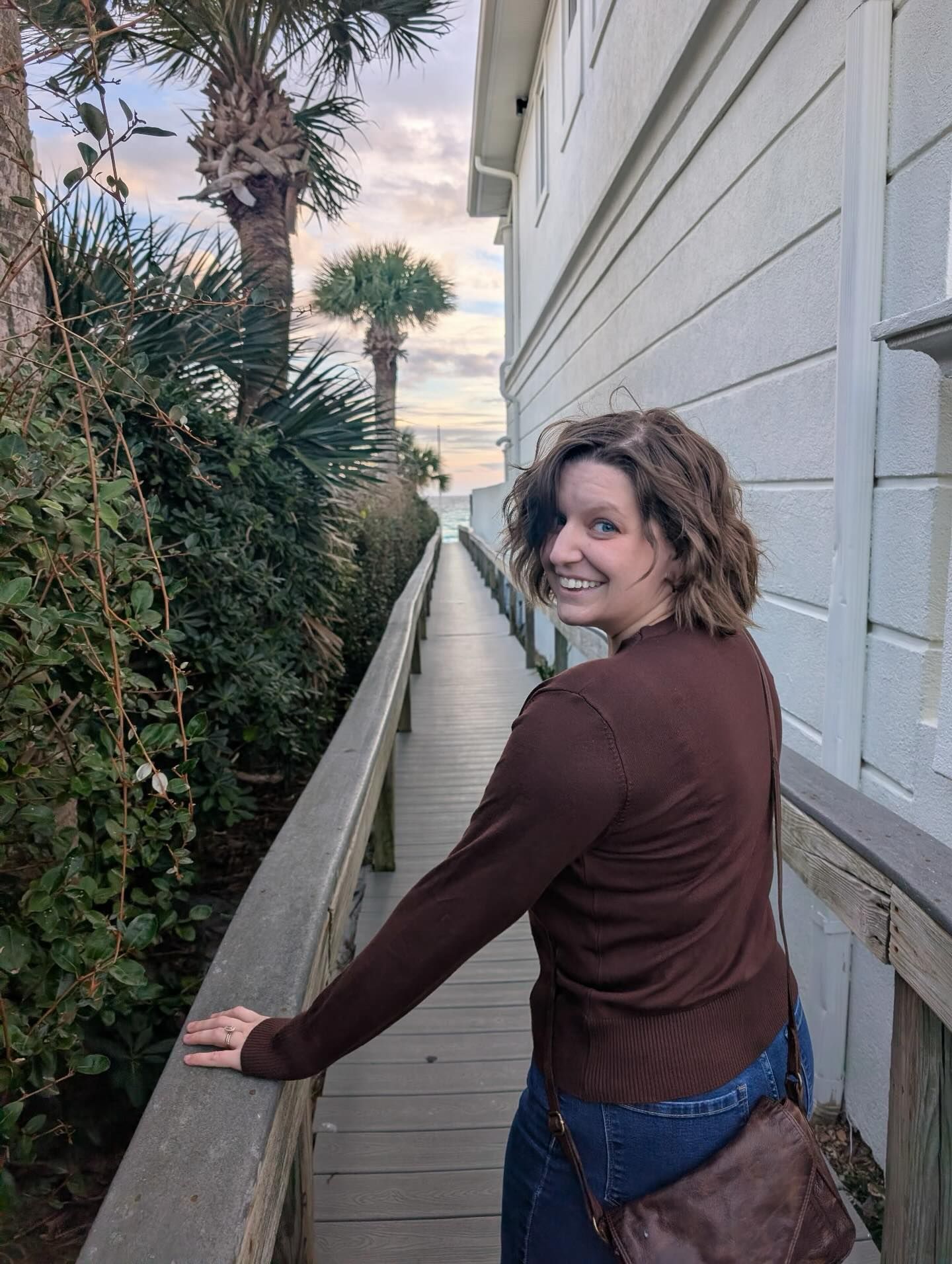 Alaynna Meyer Media in brown top smiles on a narrow boardwalk beside a beach house, with palm trees and sunset sky behind her.