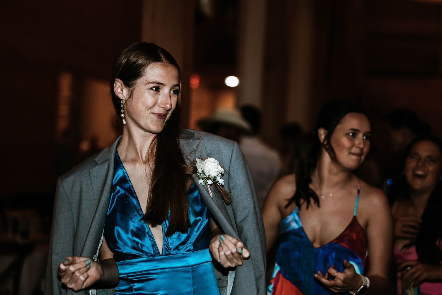 Woman in gray blazer and blue dress walks confidently at formal event, with guests in colorful attire in background.