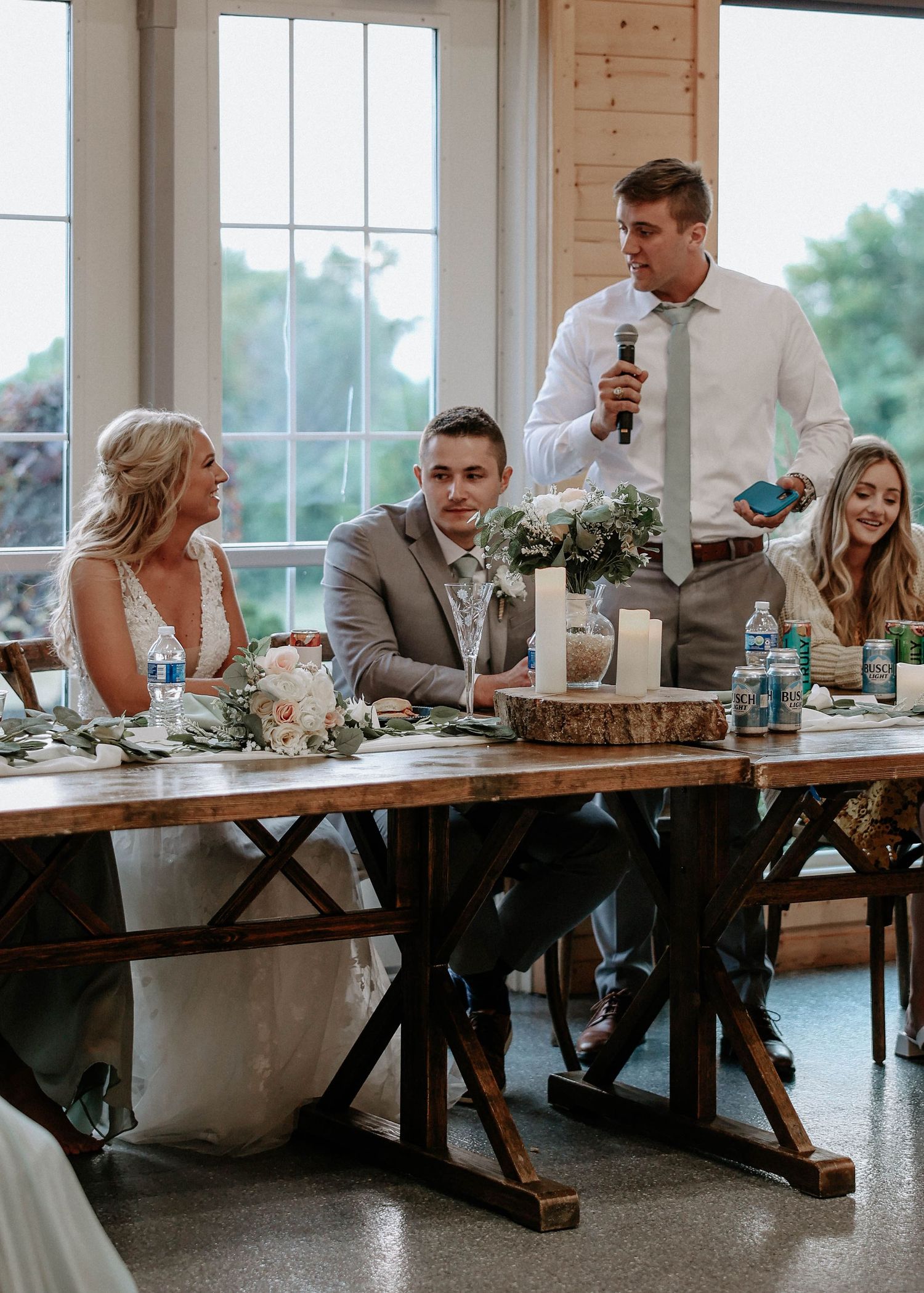 Best man delivers a heartfelt wedding speech at a rustic barn reception while bride and groom listen at decorated head table.