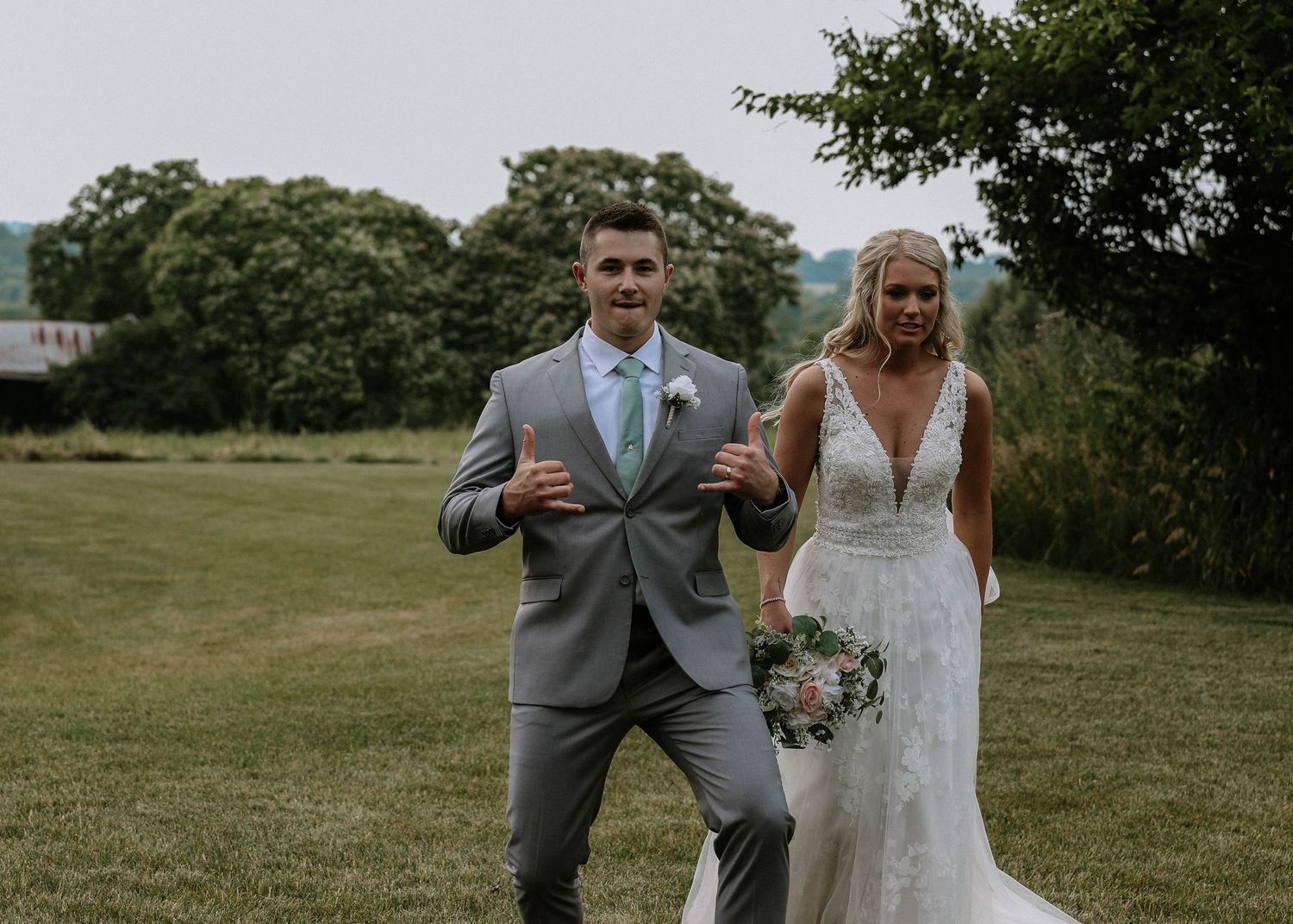Smiling groom in gray suit and bride in lace wedding dress walking outdoors on green lawn surrounded by trees.