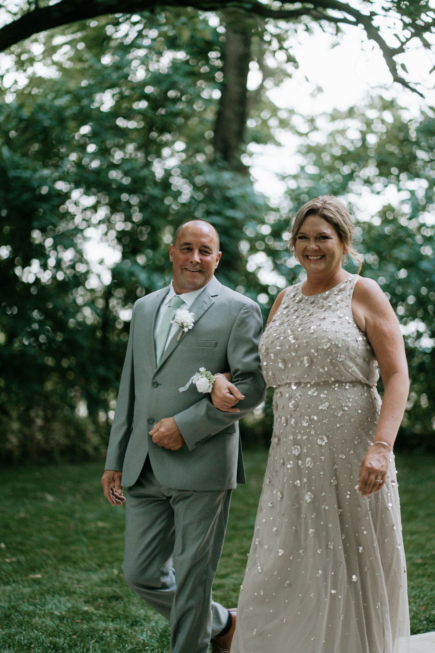 Smiling couple walking down the outdoor wedding aisle, man in gray suit, woman in beaded gown.