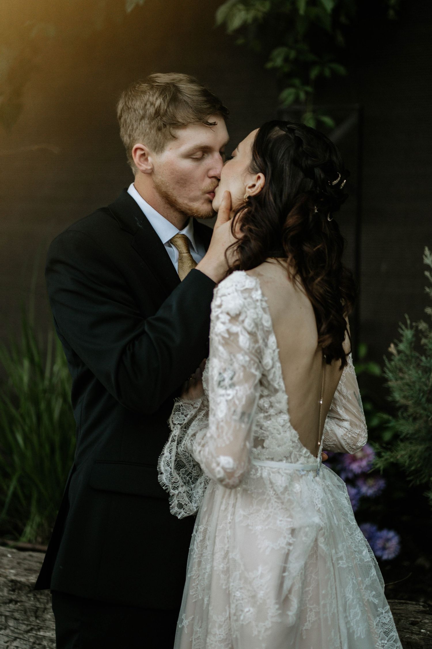 A bride and groom share a romantic kiss, she in a lace backless gown, he in a dark suit with gold tie.