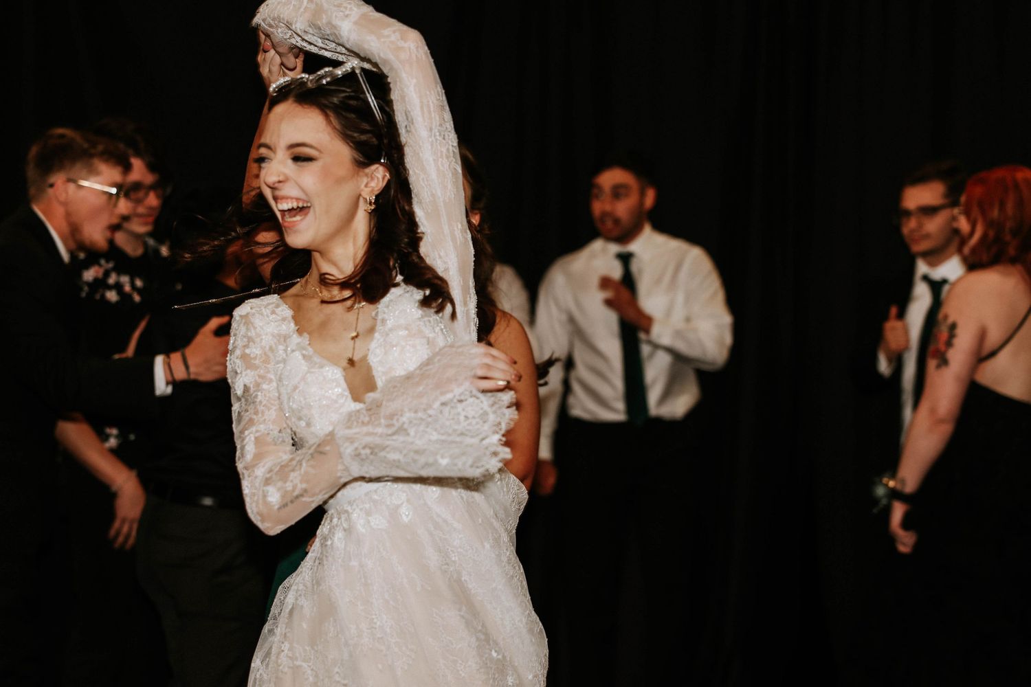 Bride in white lace gown laughing and dancing joyfully at wedding reception with guests in background.
