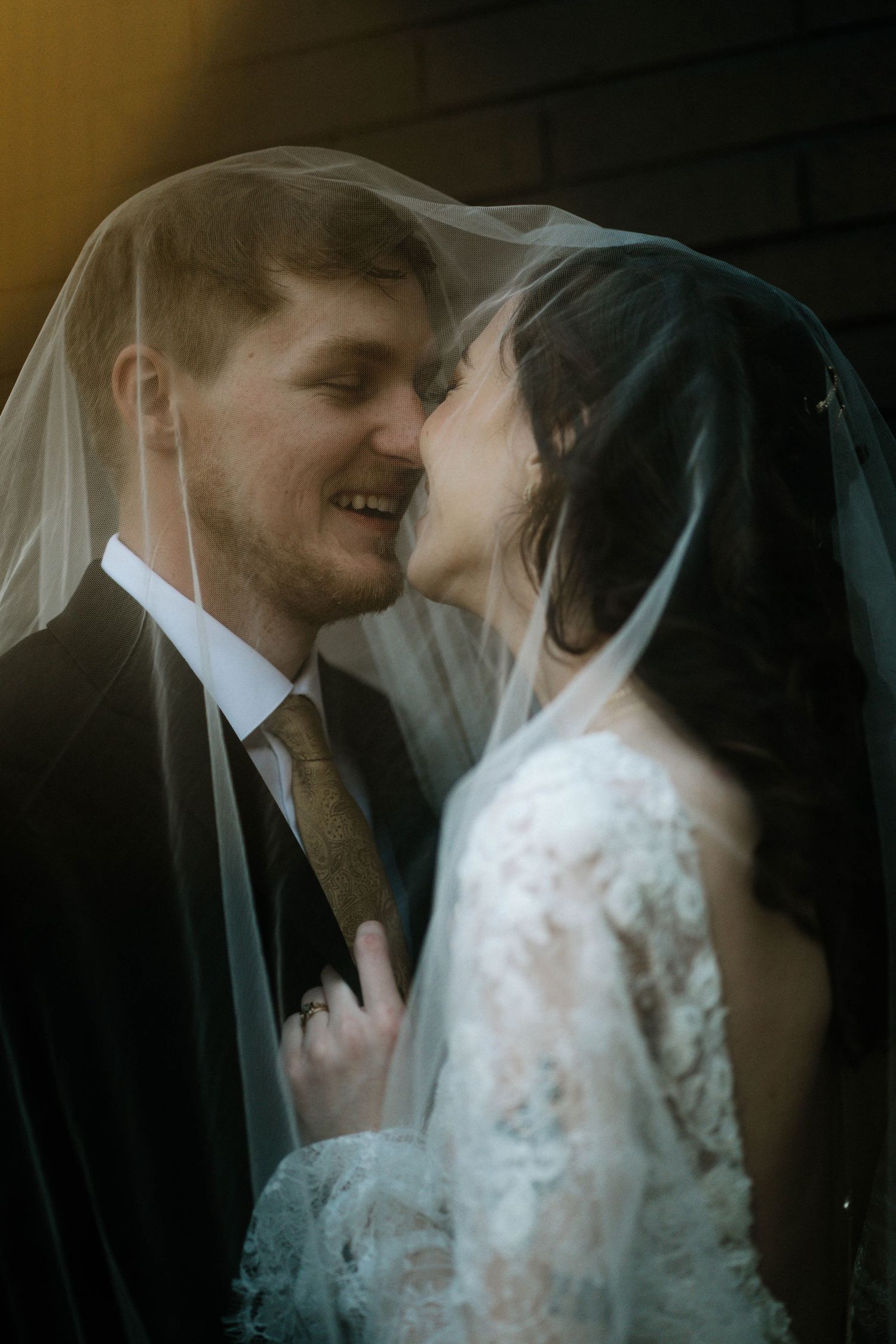 Bride and groom share a romantic moment under a sheer veil, smiling closely together in wedding attire.