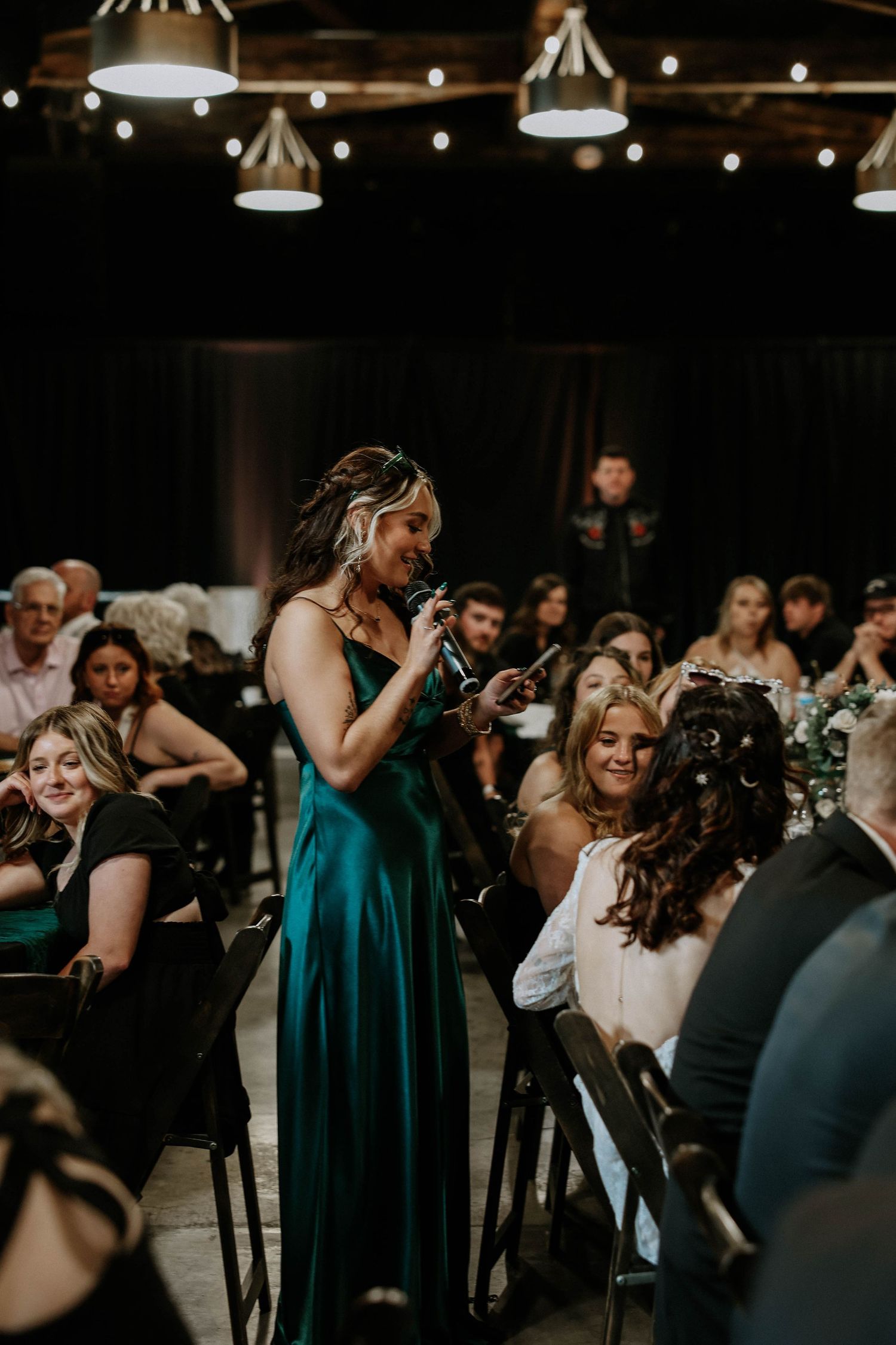 A woman in an emerald green satin gown gives a speech with a microphone at an elegant indoor wedding reception.