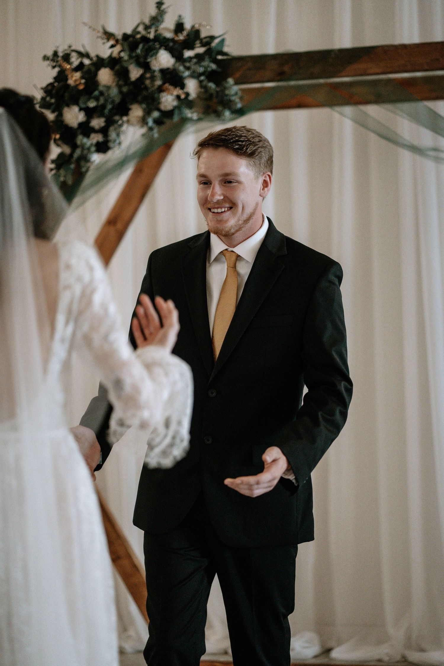 Groom in black suit and gold tie smiling at bride during indoor wedding ceremony with floral arch backdrop.