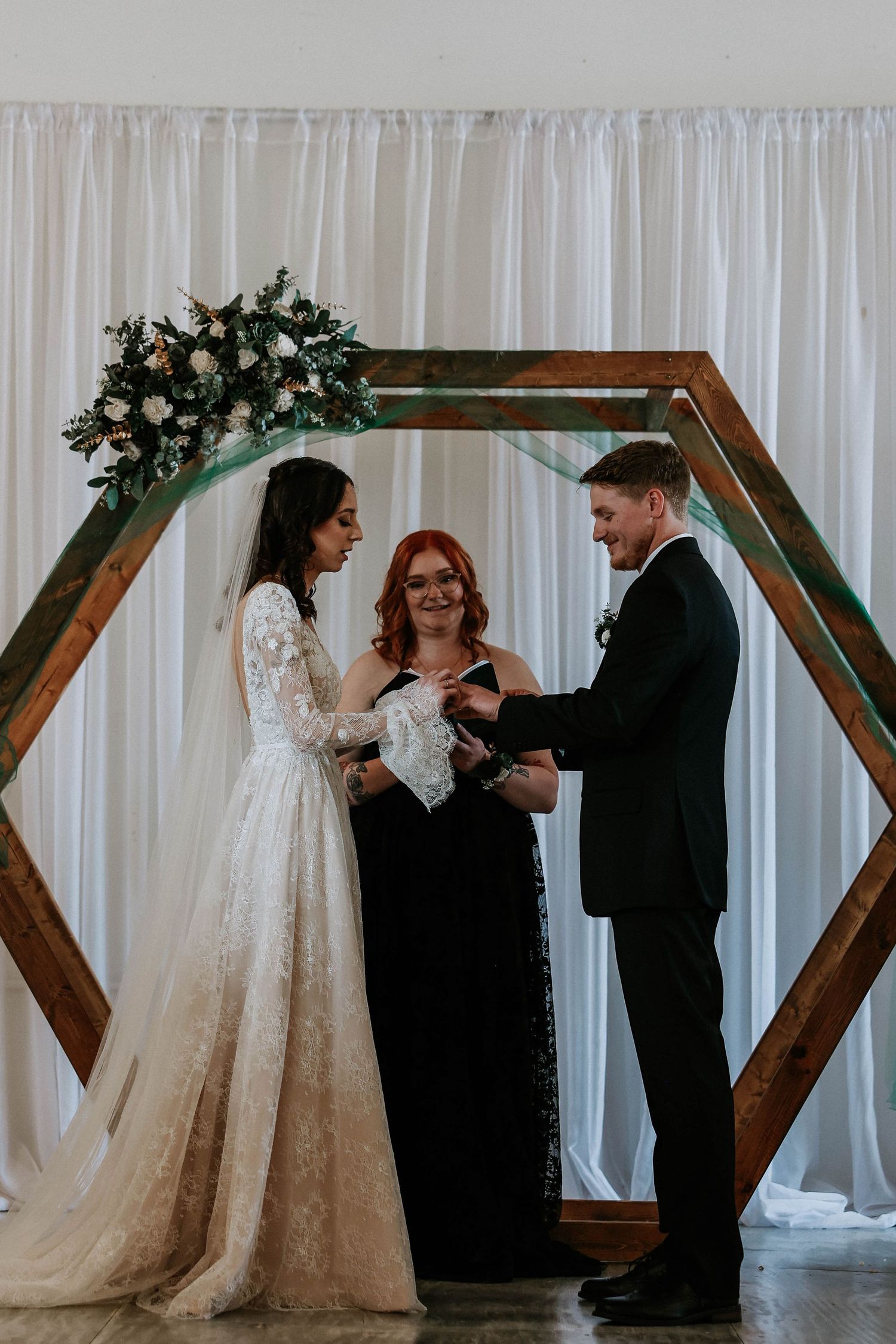 Bride and groom exchange vows before officiant under wooden hexagonal arch with floral decor and white drape backdrop.