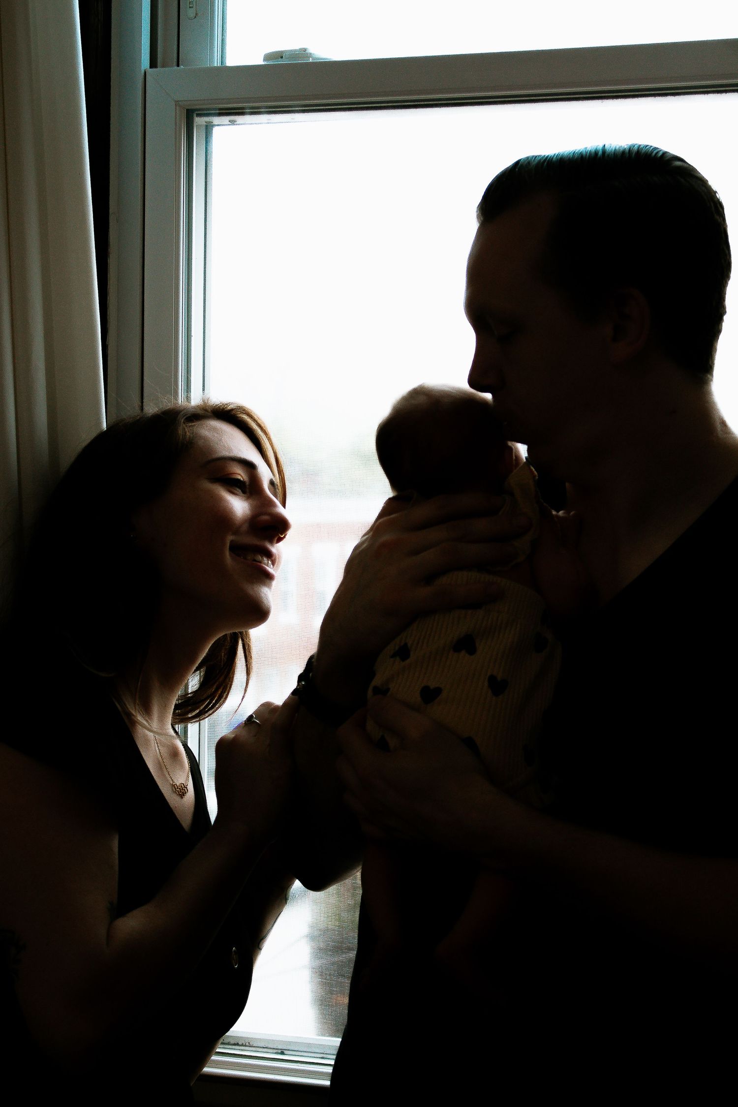 Silhouette of smiling parents holding newborn baby by window in tender family moment at Mill Street Studios in Cincinnati, Ohio.