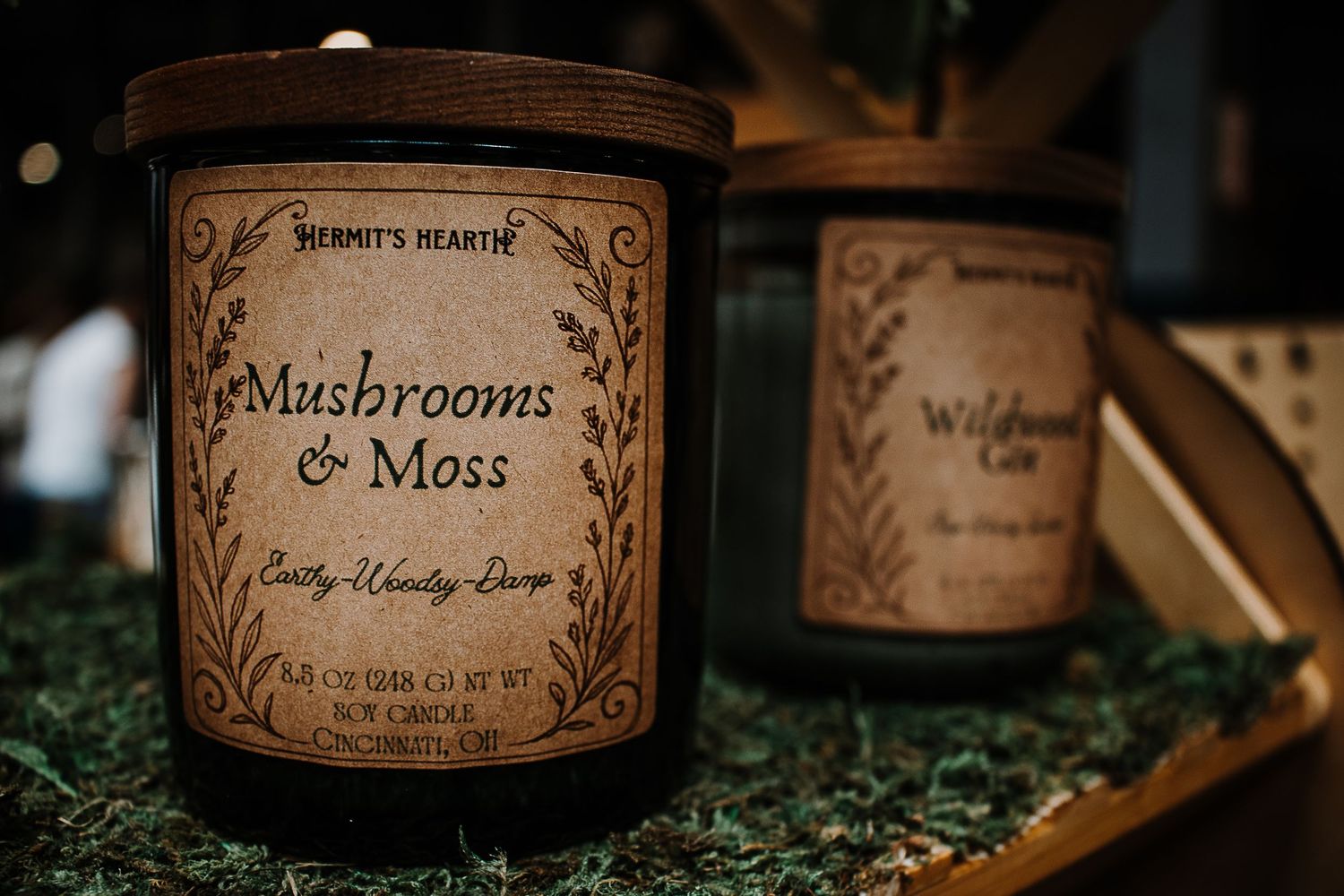 Hermit's Hearth Mushrooms & Moss soy candle in dark jar with wooden lid, beside Wildflower candle on mossy surface.