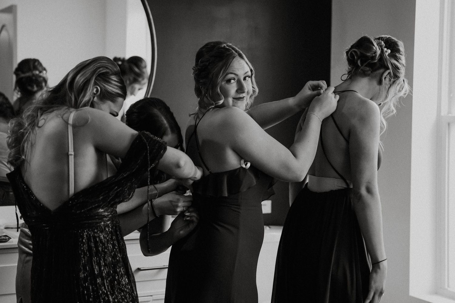 Bridesmaids helping bride zip up her elegant gown in black and white wedding preparation photo.