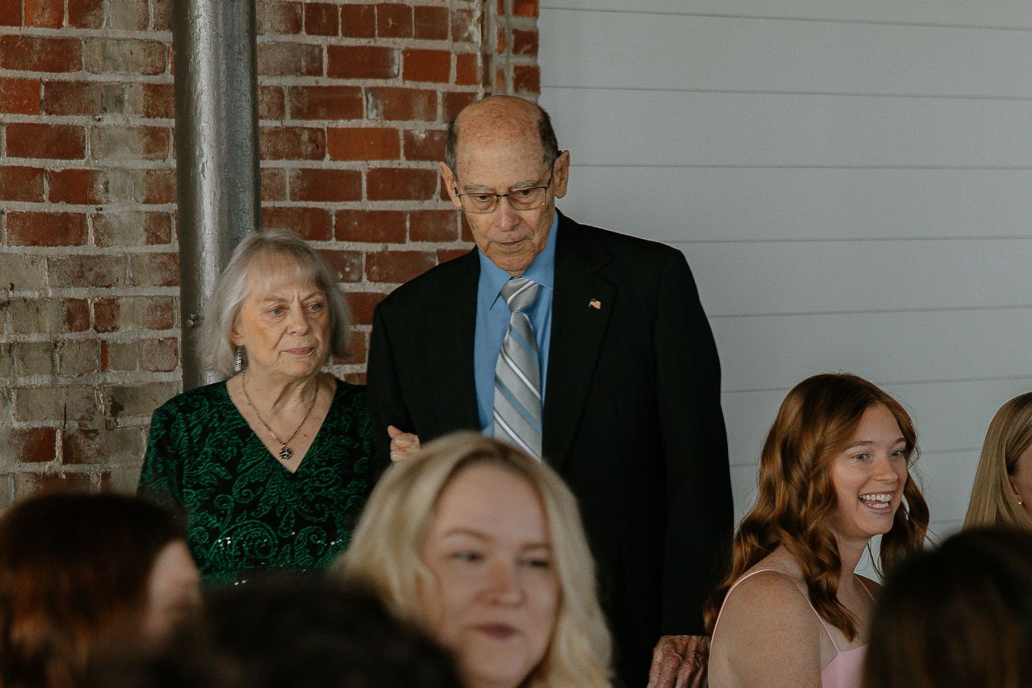 Elderly couple walking together at a wedding ceremony beside exposed brick wall.