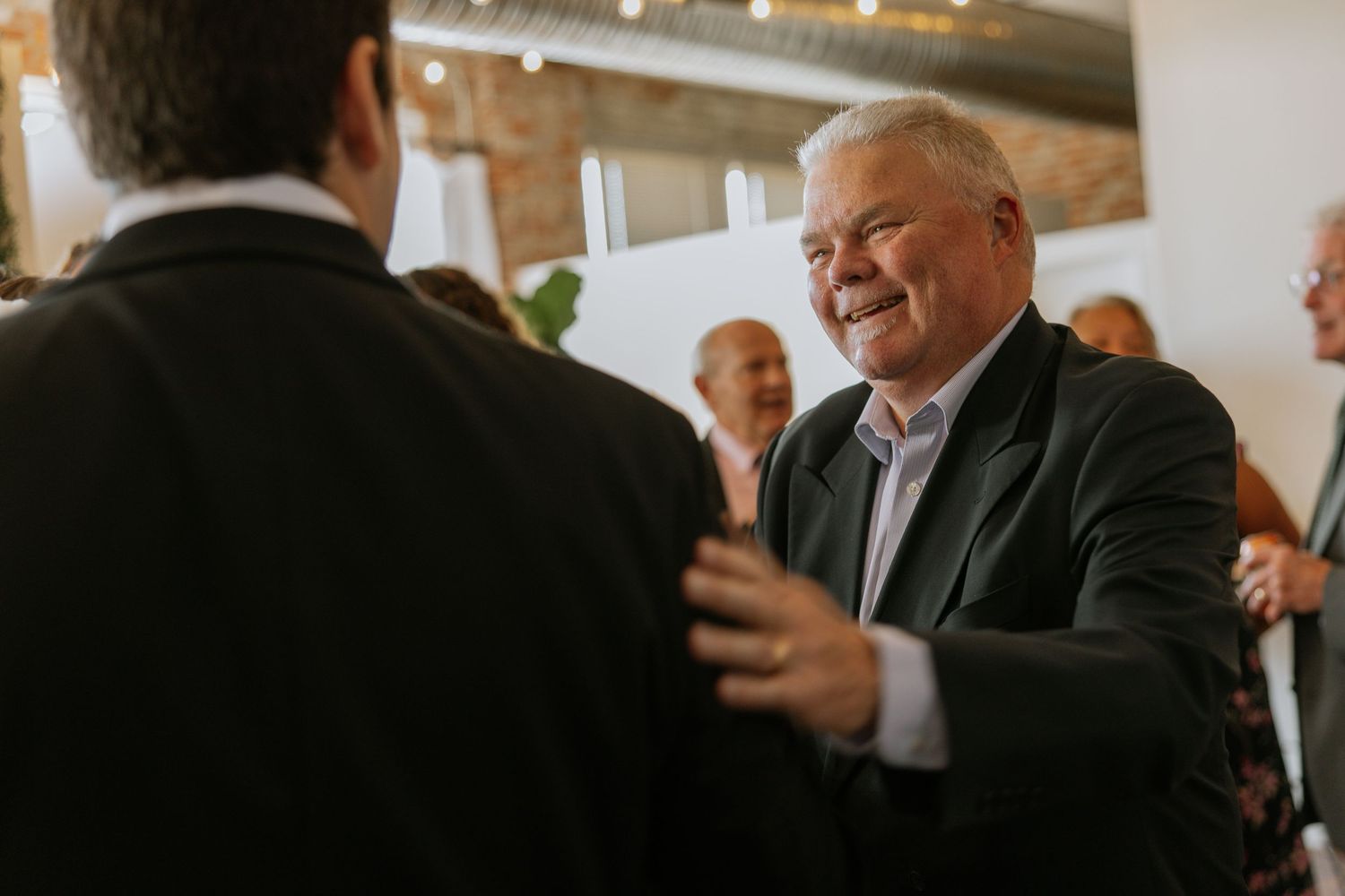 Elderly man in dark suit smiling warmly while greeting guests at an elegant wedding reception.