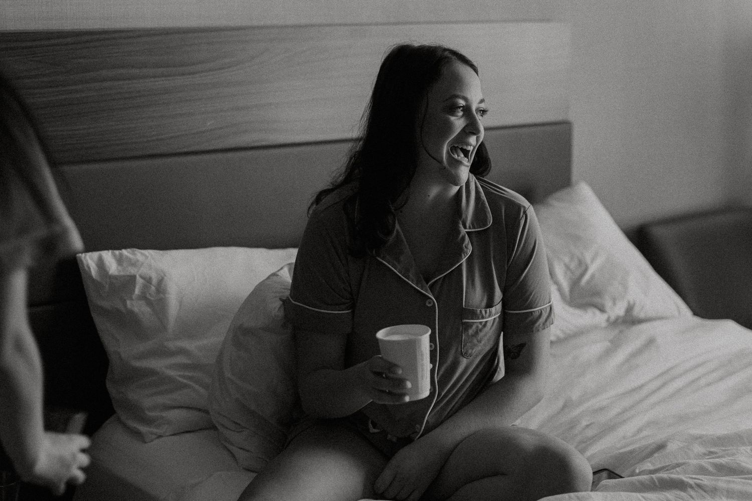 Woman in pajamas laughing while holding a coffee mug sitting on a hotel bed in black and white photo.