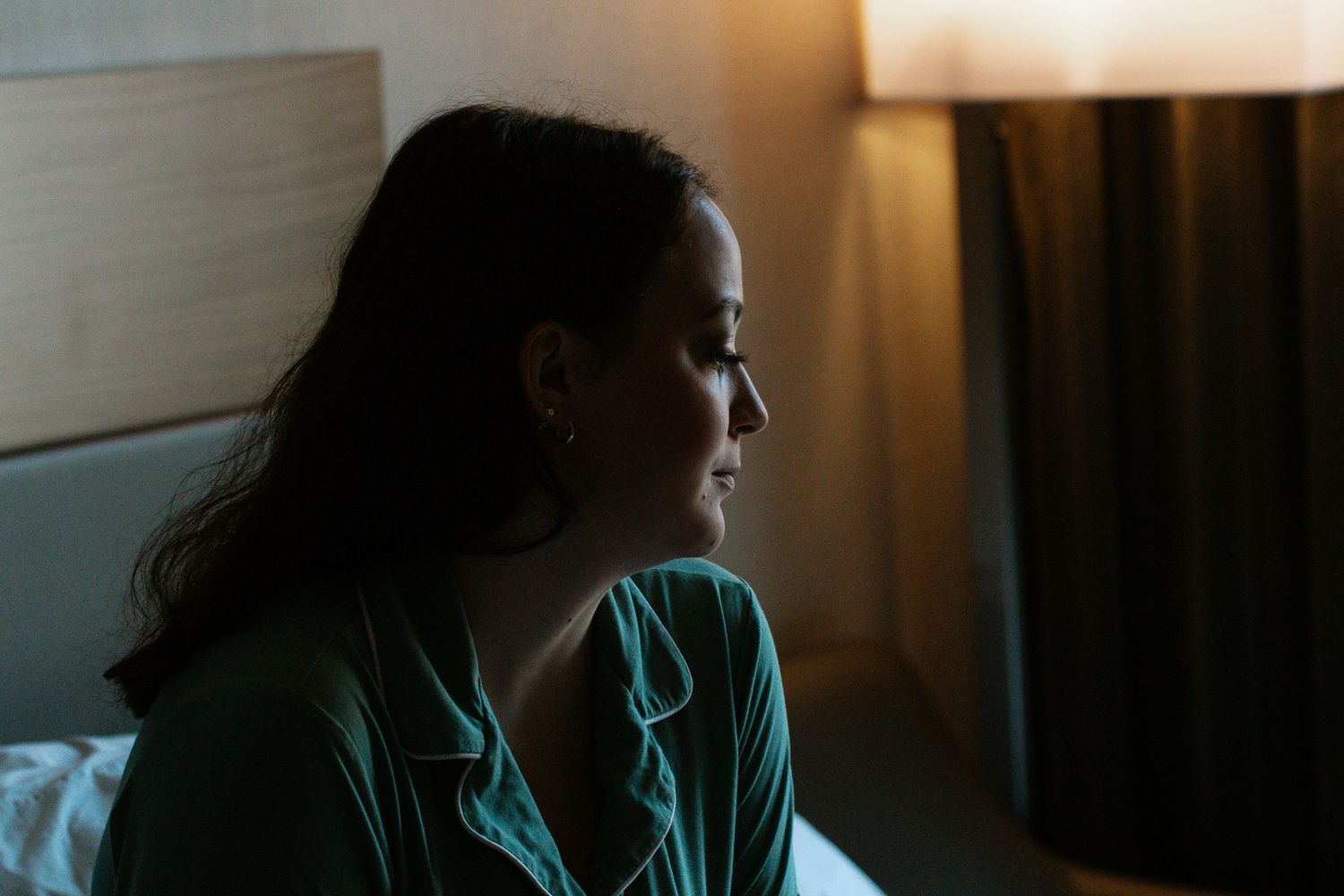 Bride in green satin pajamas sits on hotel bed in profile, illuminated by warm bedside lamp light.