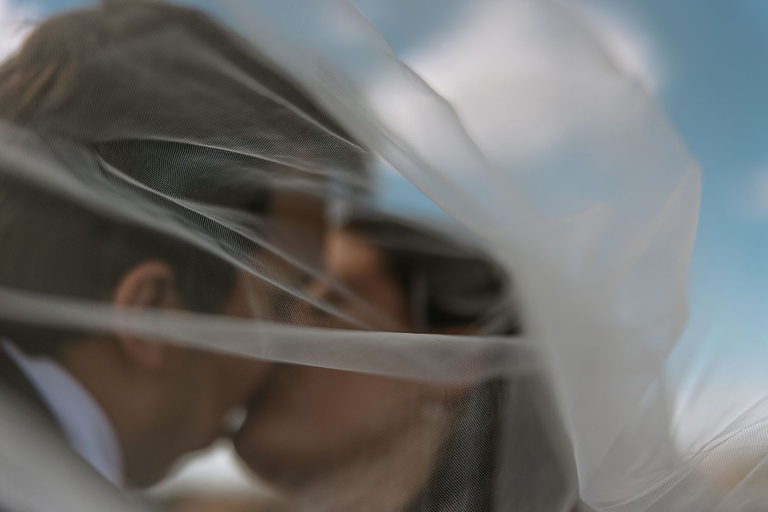 Dreamy artistic wedding photo of couple kissing seen through layers of flowing bridal veil fabric.