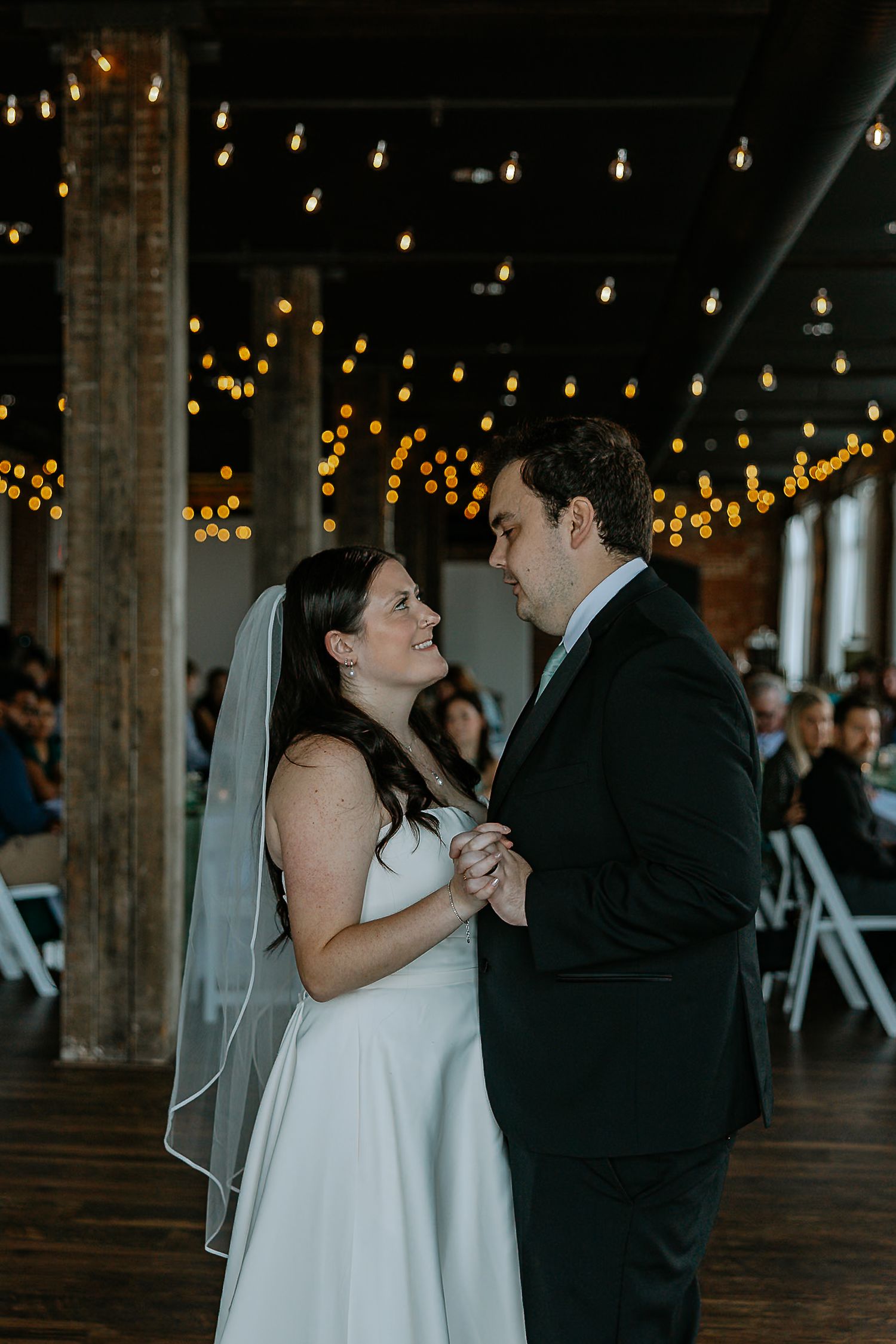 Bride and groom share their first dance, gazing into each other's eyes under warm string lights.