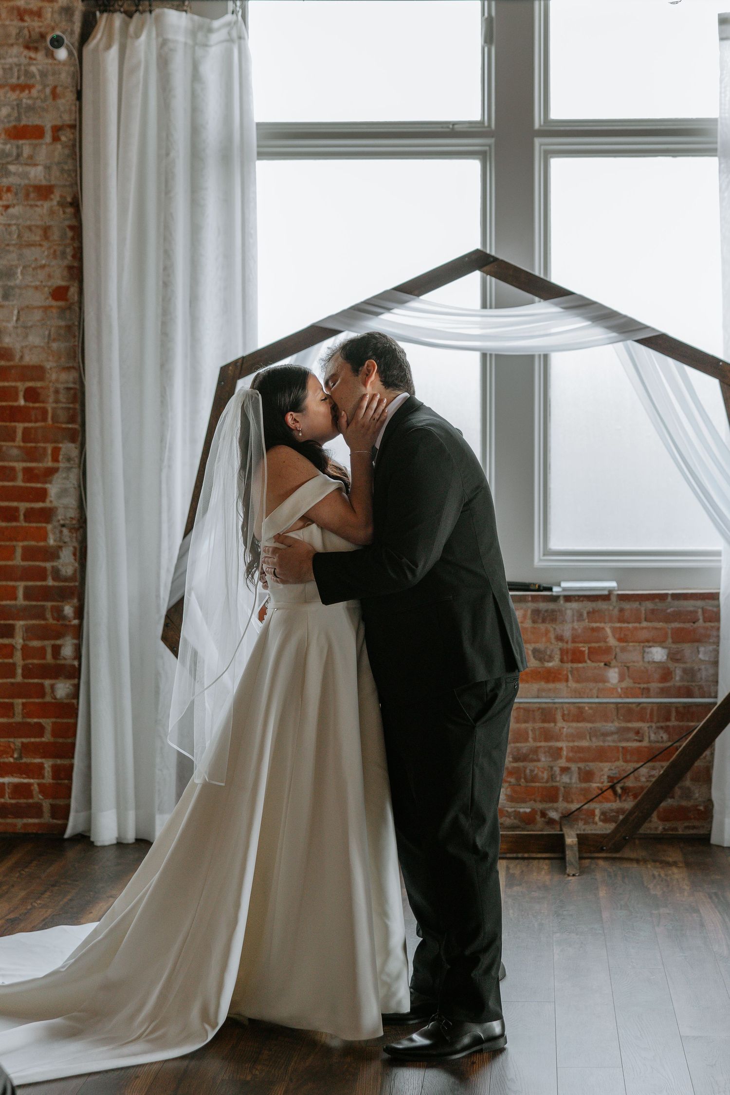 Bride and groom sharing first kiss under wooden arch at wedding ceremony.