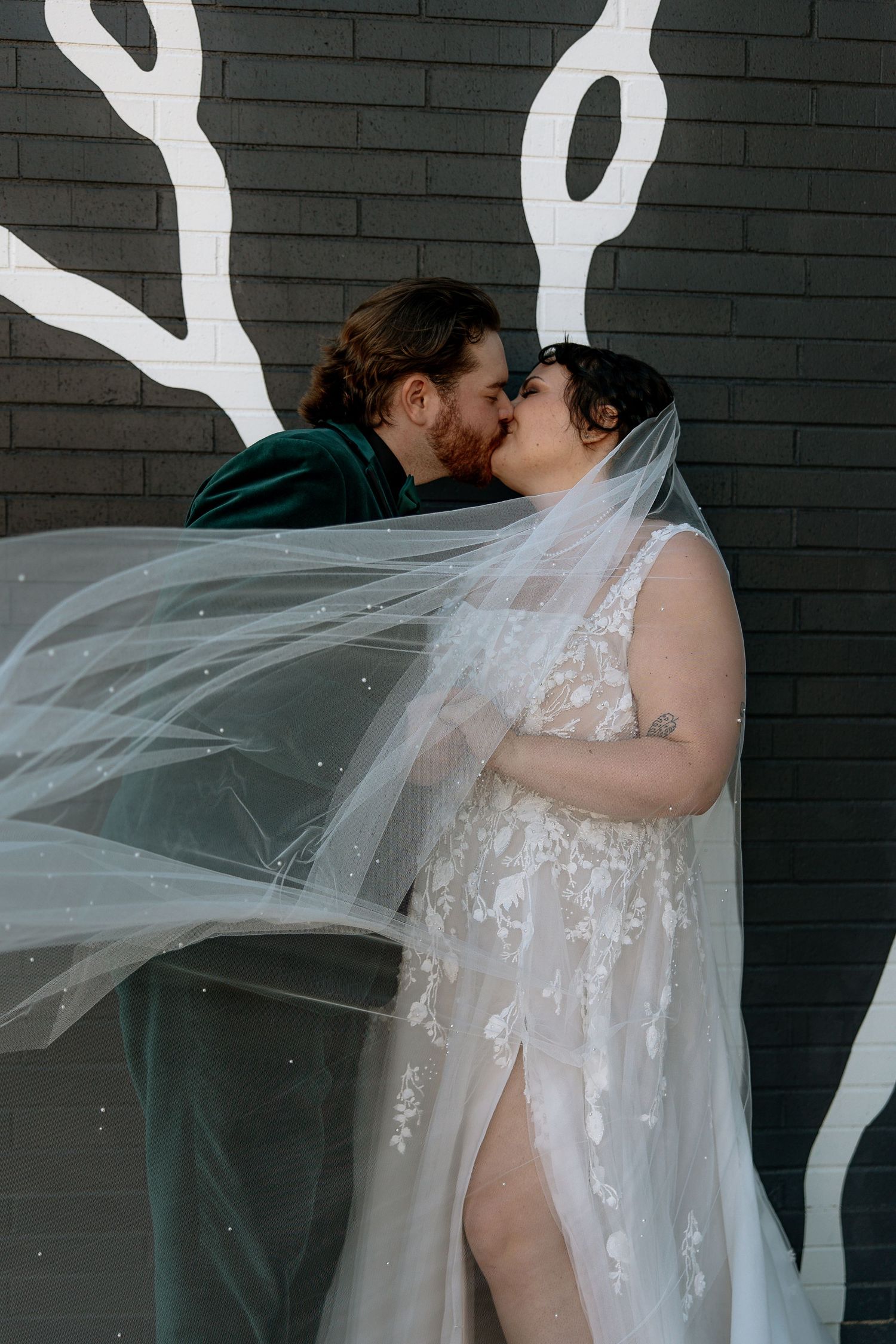 Bride and groom share kiss against dark mural wall as flowing veil billows dramatically around them.