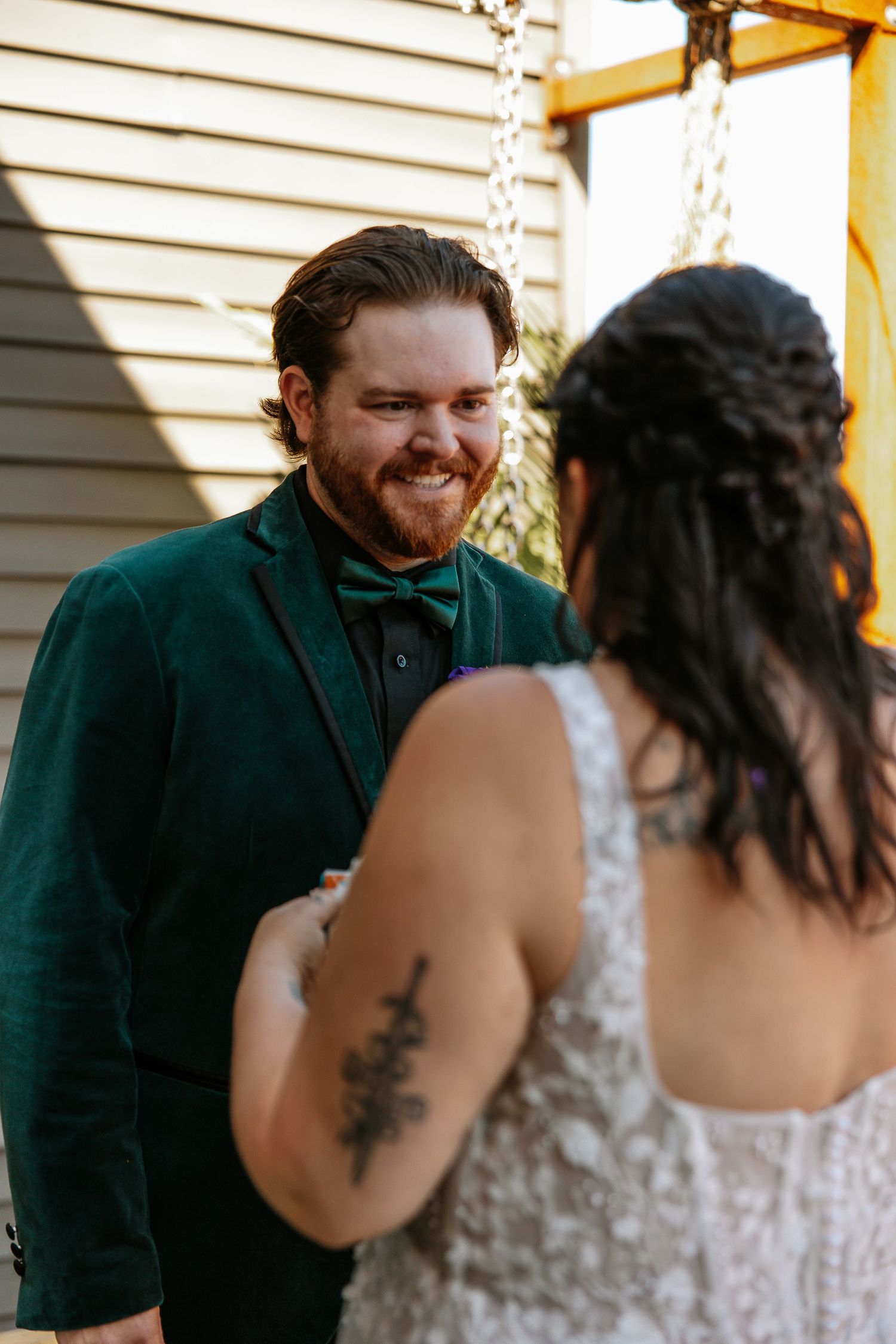 Groom in emerald velvet suit smiles joyfully at tattooed bride in floral lace wedding dress.