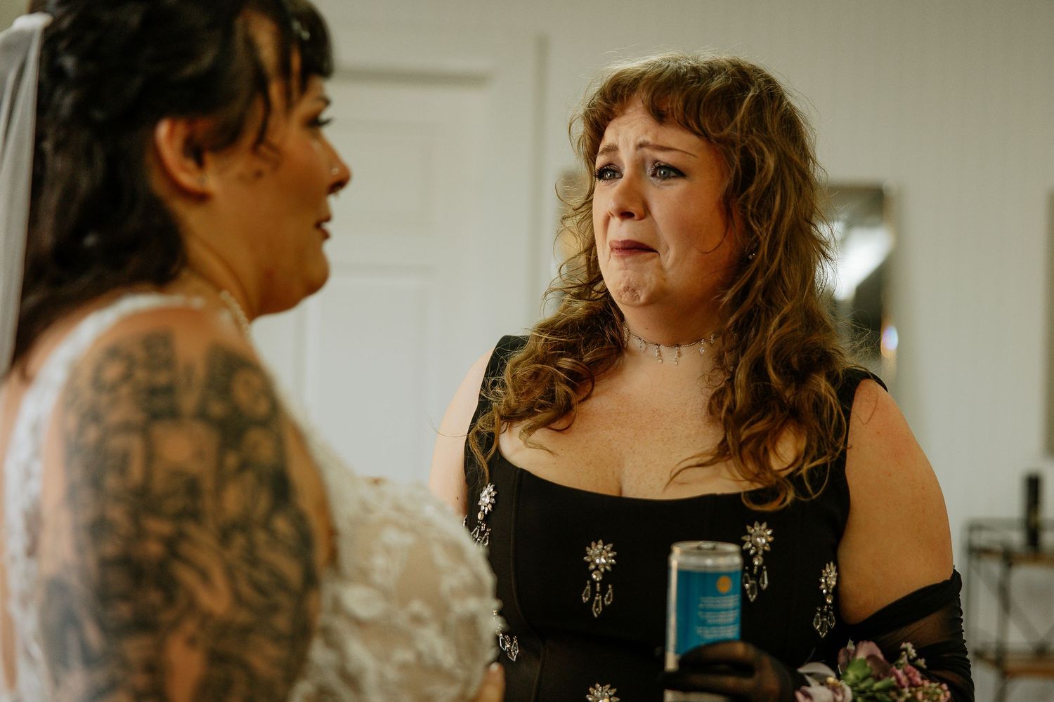 Emotional mother in black dress holding a drink while speaking with tattooed bride in white wedding gown.