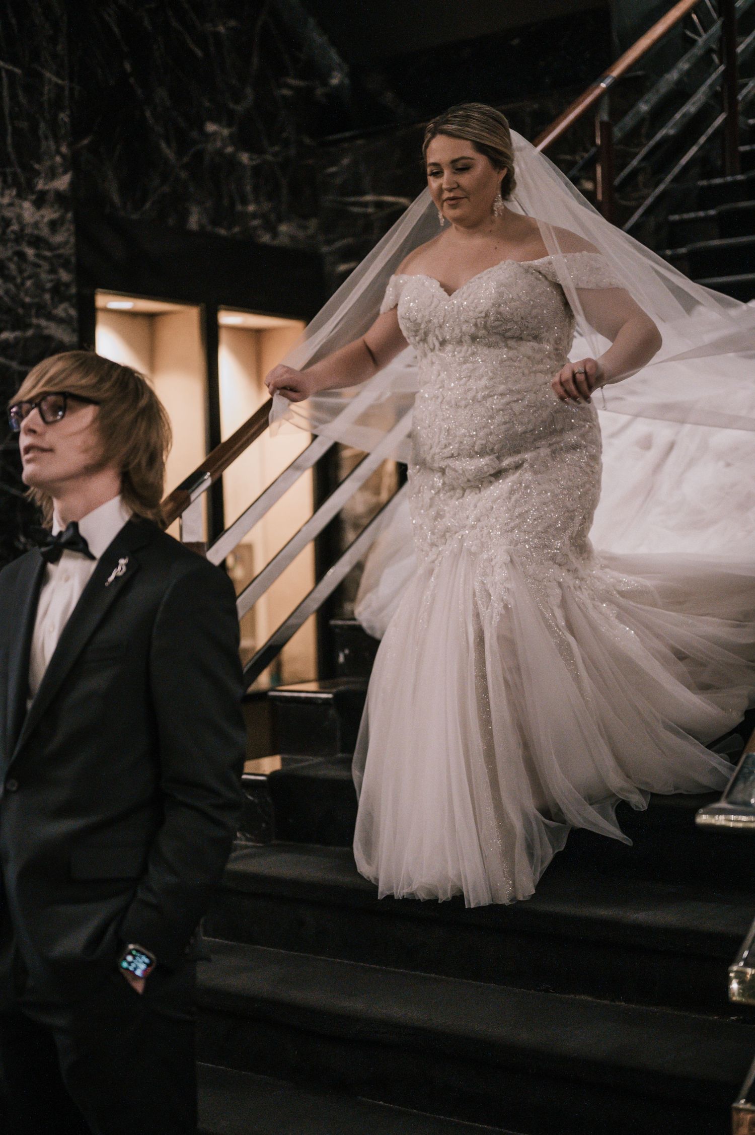 Bride in beaded mermaid gown with veil descends staircase beside young man in black tuxedo.