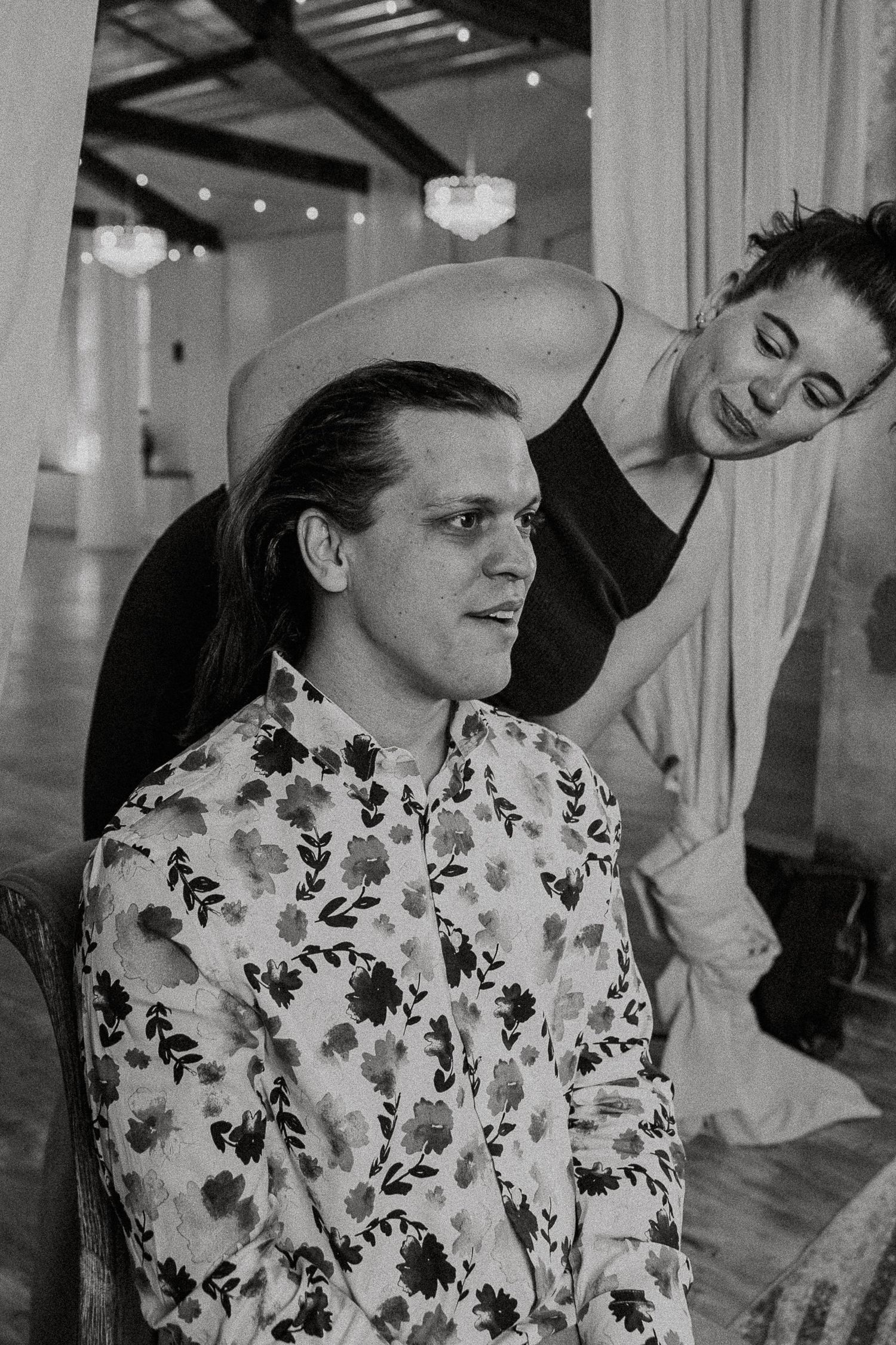 Black and white photo of man in floral shirt and hat getting hair styled.