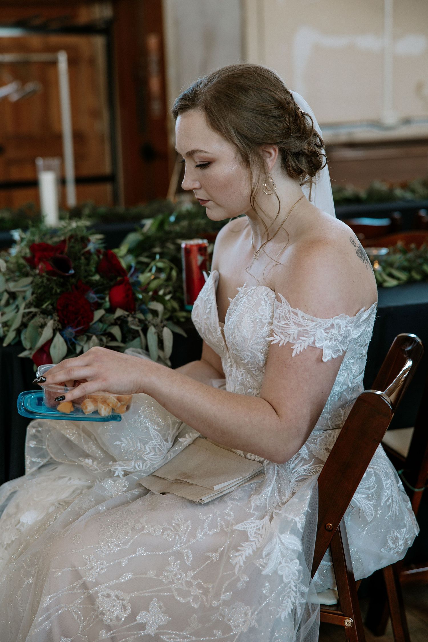 Bride in lace off-shoulder wedding gown sits at table holding a small item during reception at Urban Artifact in Cincinnati, Ohio