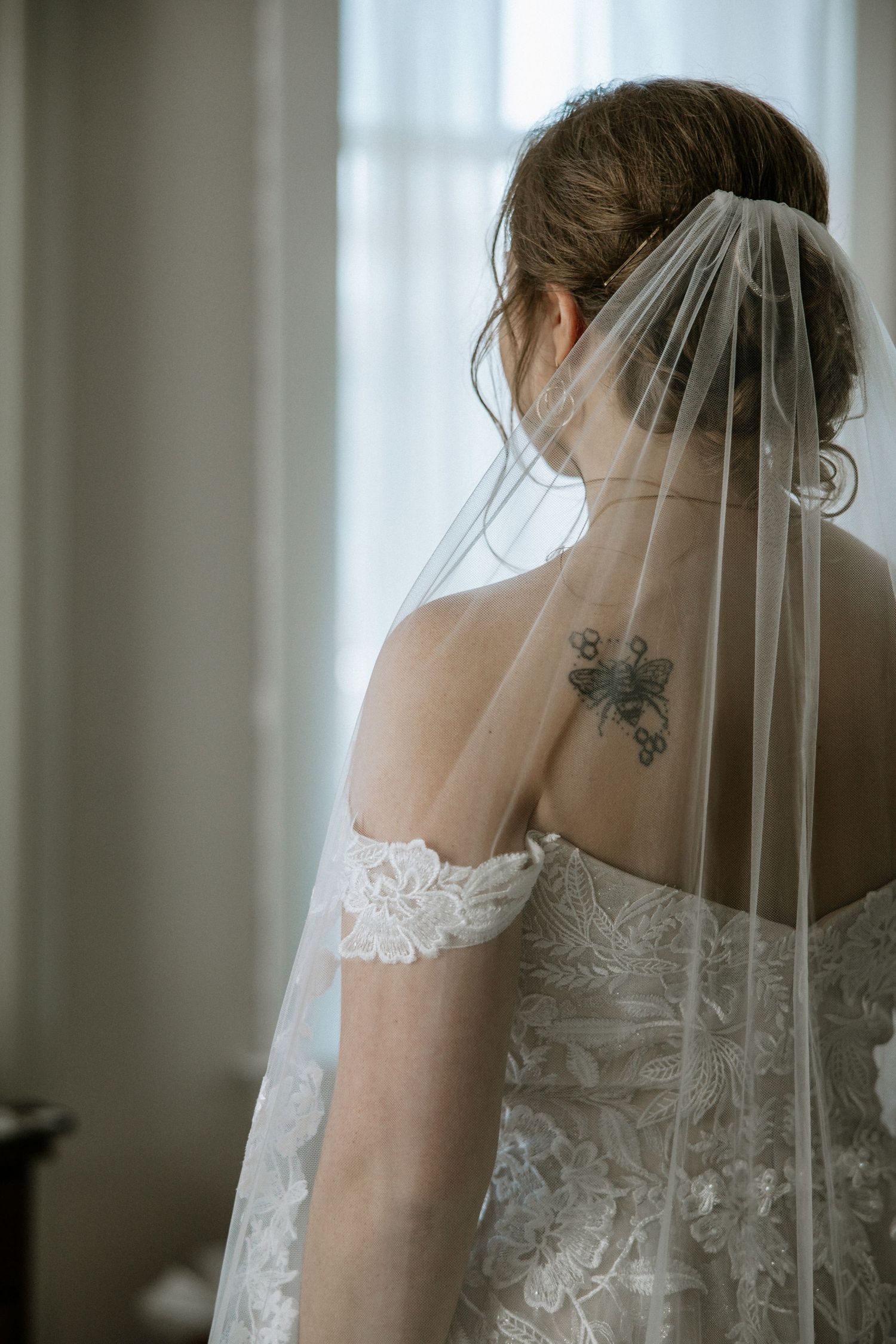 Bride with floral back tattoo wearing an off-shoulder lace gown and sheer veil by a window at Urban Artifact in Cincinnati, Ohio