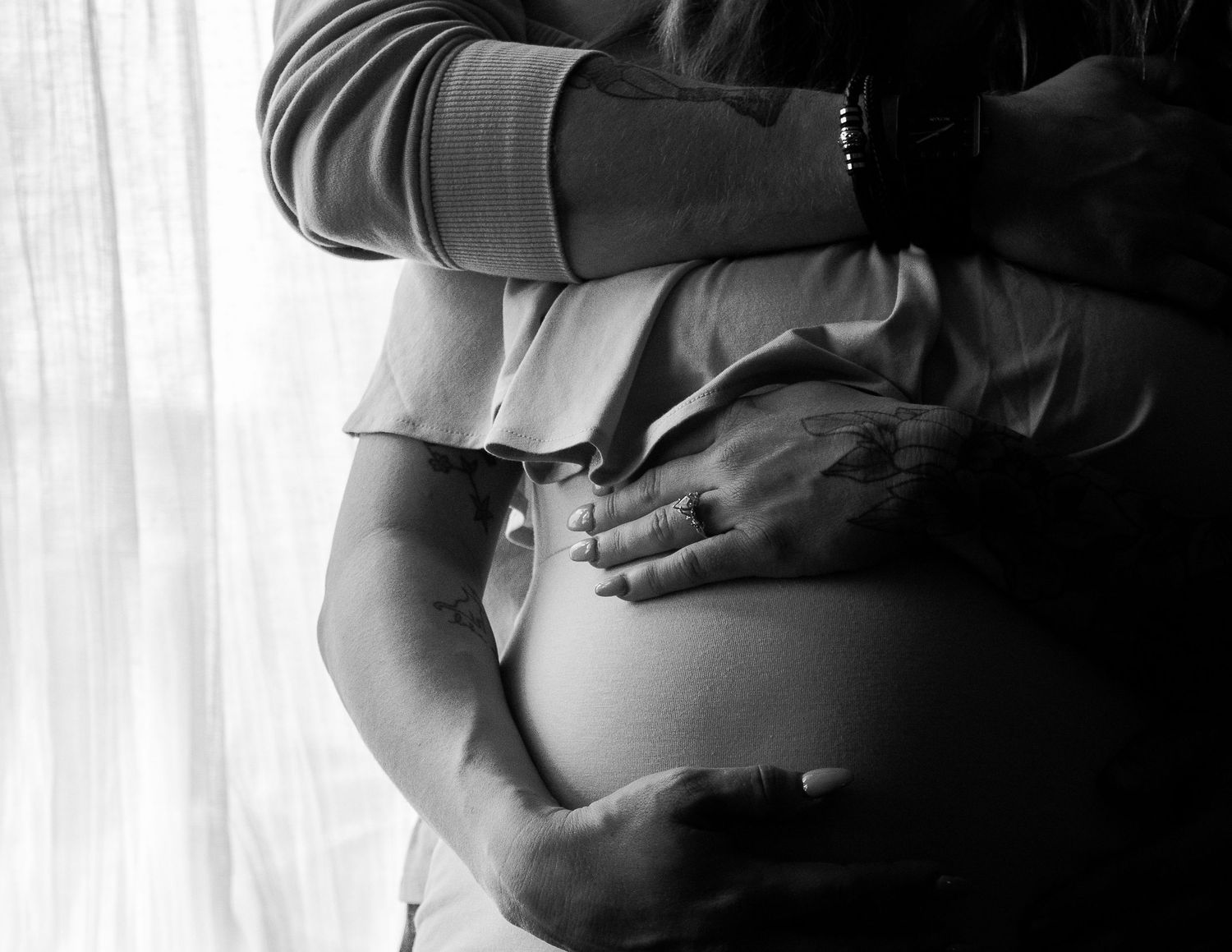 Partner embracing pregnant woman from behind near sheer curtained window at Mill Street Studios in Cincinnati, Ohio.