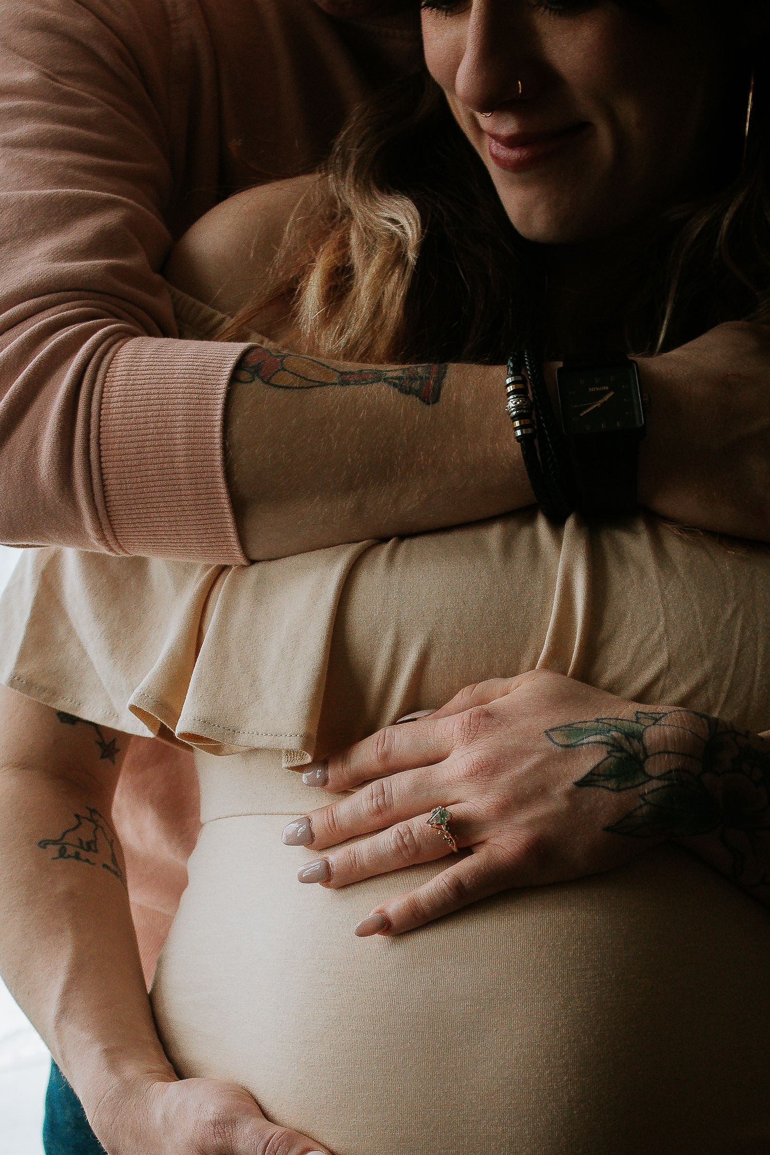 Partner embracing tattooed pregnant woman while holding baby bump in Cincinnati, Ohio.