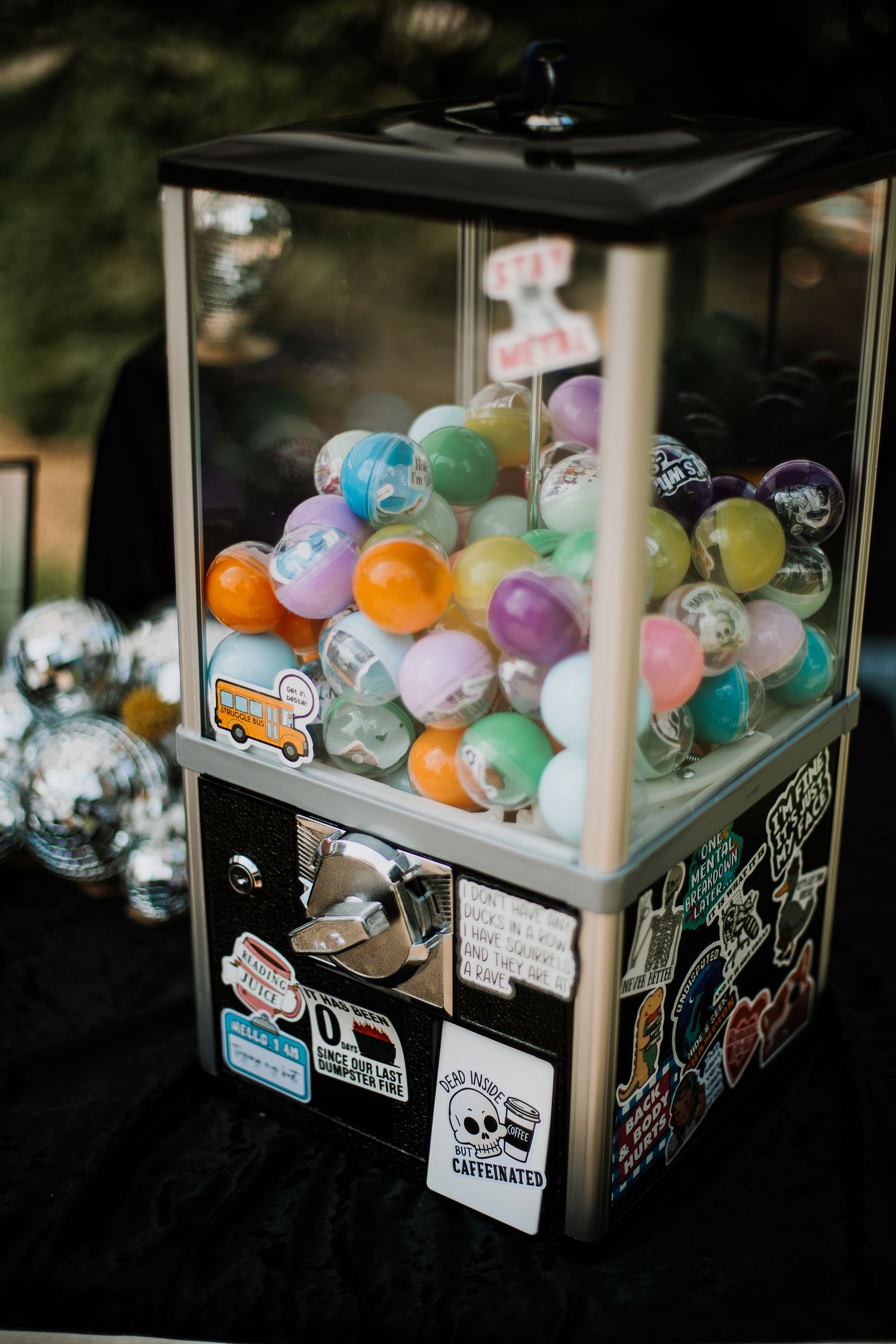 Colorful gumball machine covered in stickers filled with pastel capsule toys, displayed on a black table with disco balls.