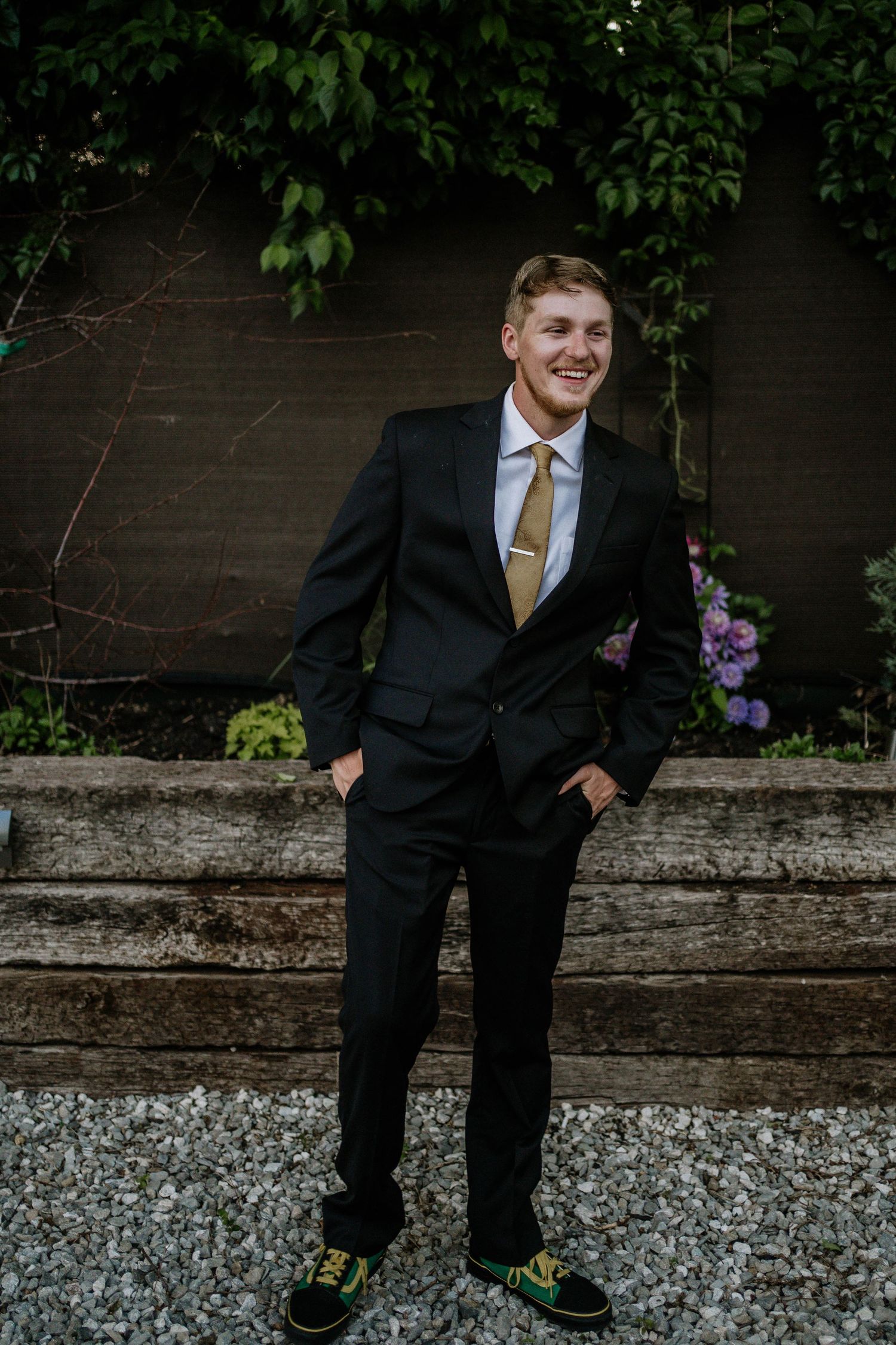 Young man in black suit with gold tie smiling outdoors, standing on gravel path with green foliage background.