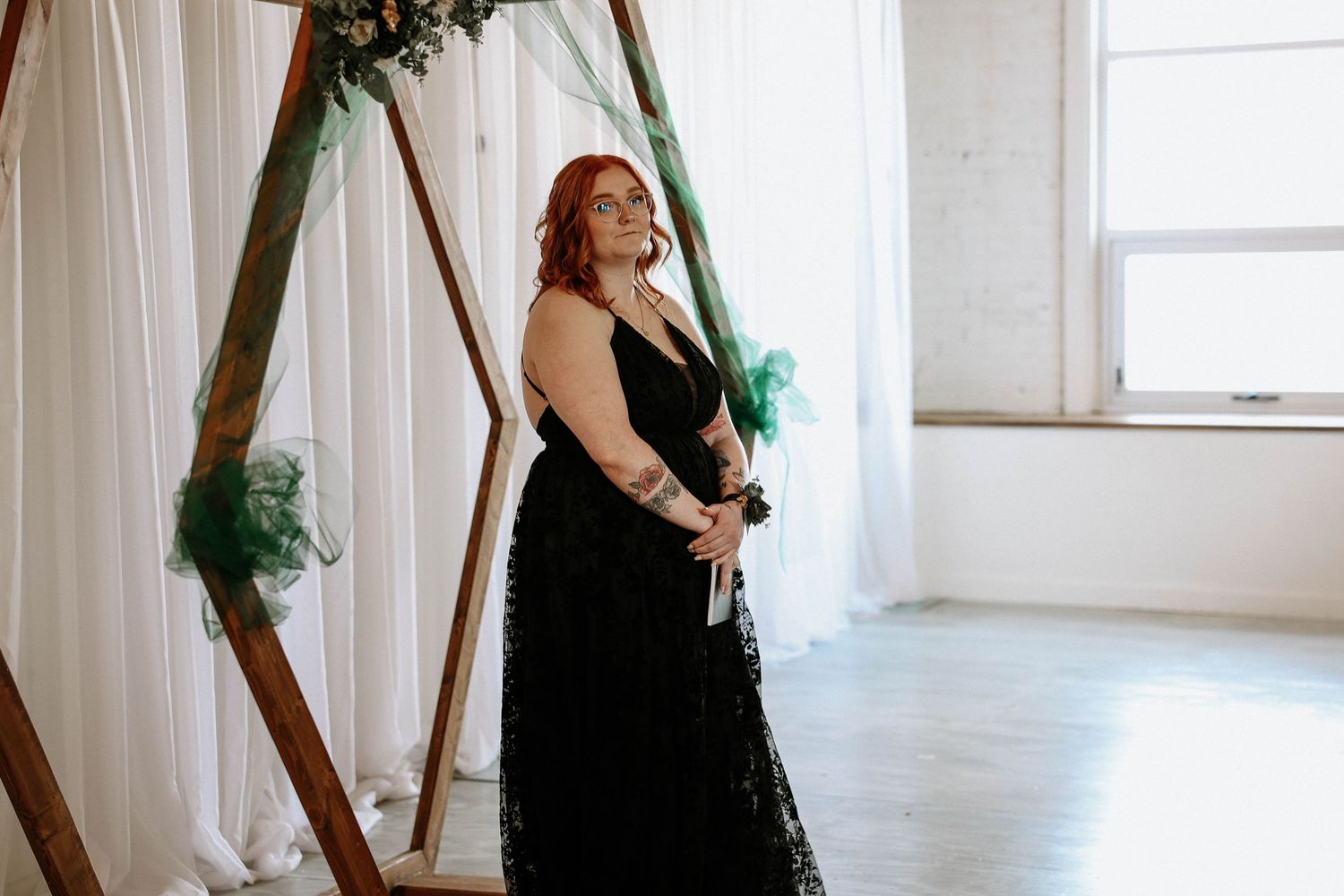 Woman in black lace gown stands before wooden geometric arch with green floral accents in bright white wedding venue.