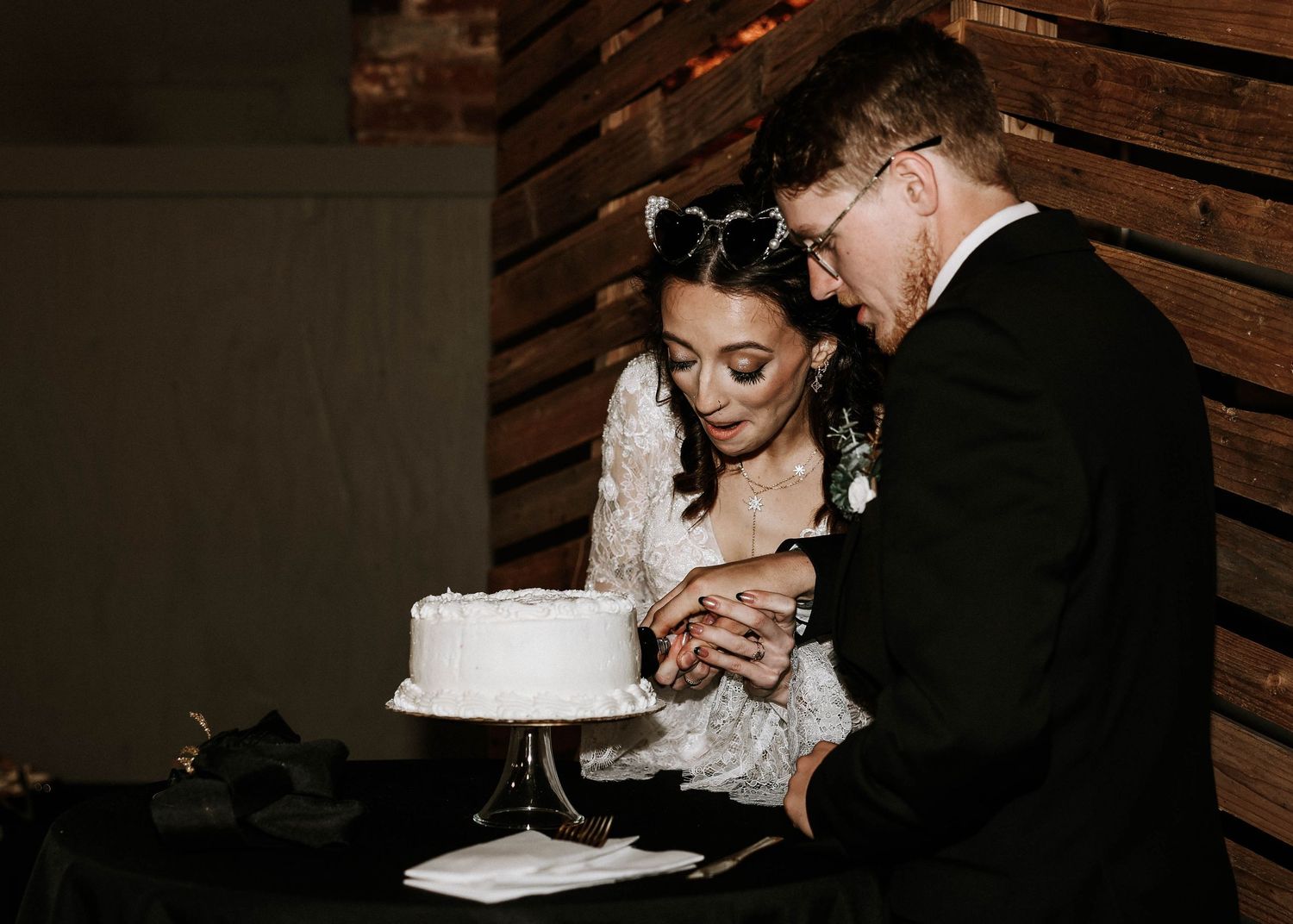 Bride and groom cutting white wedding cake together in romantic dark reception venue.