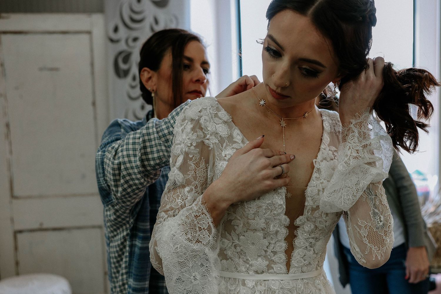 Bride in lace wedding gown being helped by bridesmaid adjusting dress and jewelry before wedding ceremony.