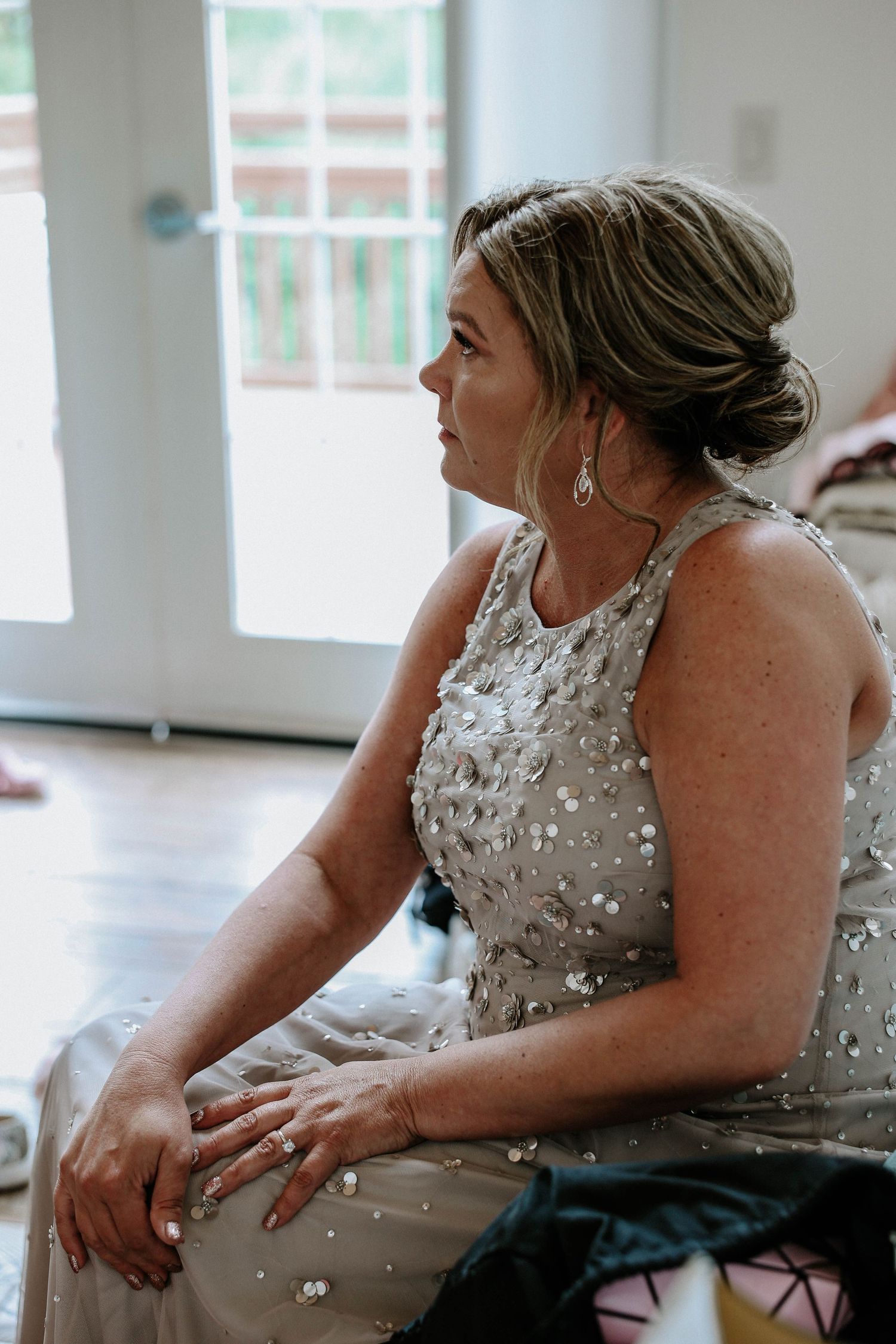 Elegant woman in silver sequined gown with updo hairstyle sitting gracefully at a wedding venue.