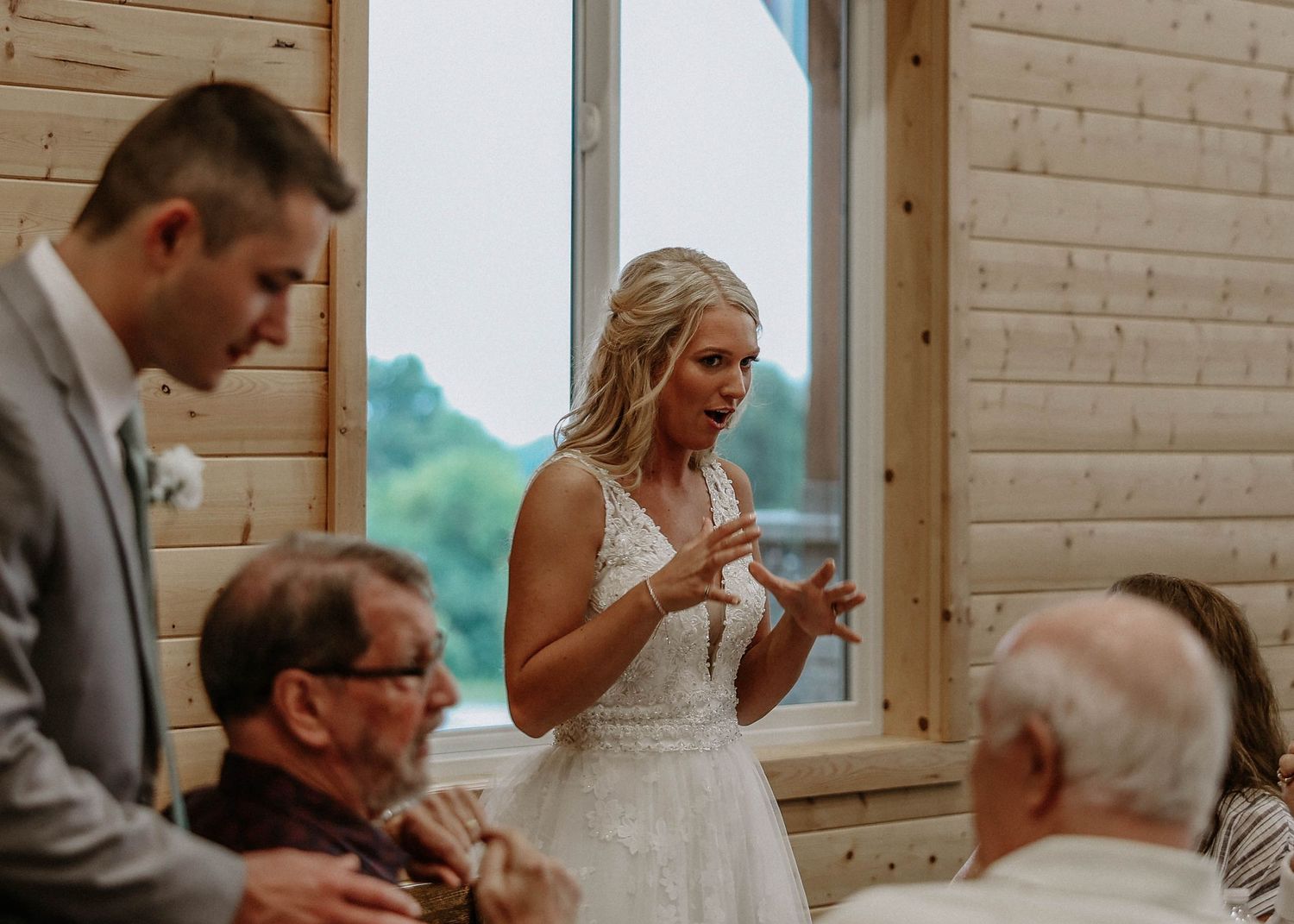 Bride in white lace gown speaking animatedly to seated wedding guests in a rustic log cabin venue.