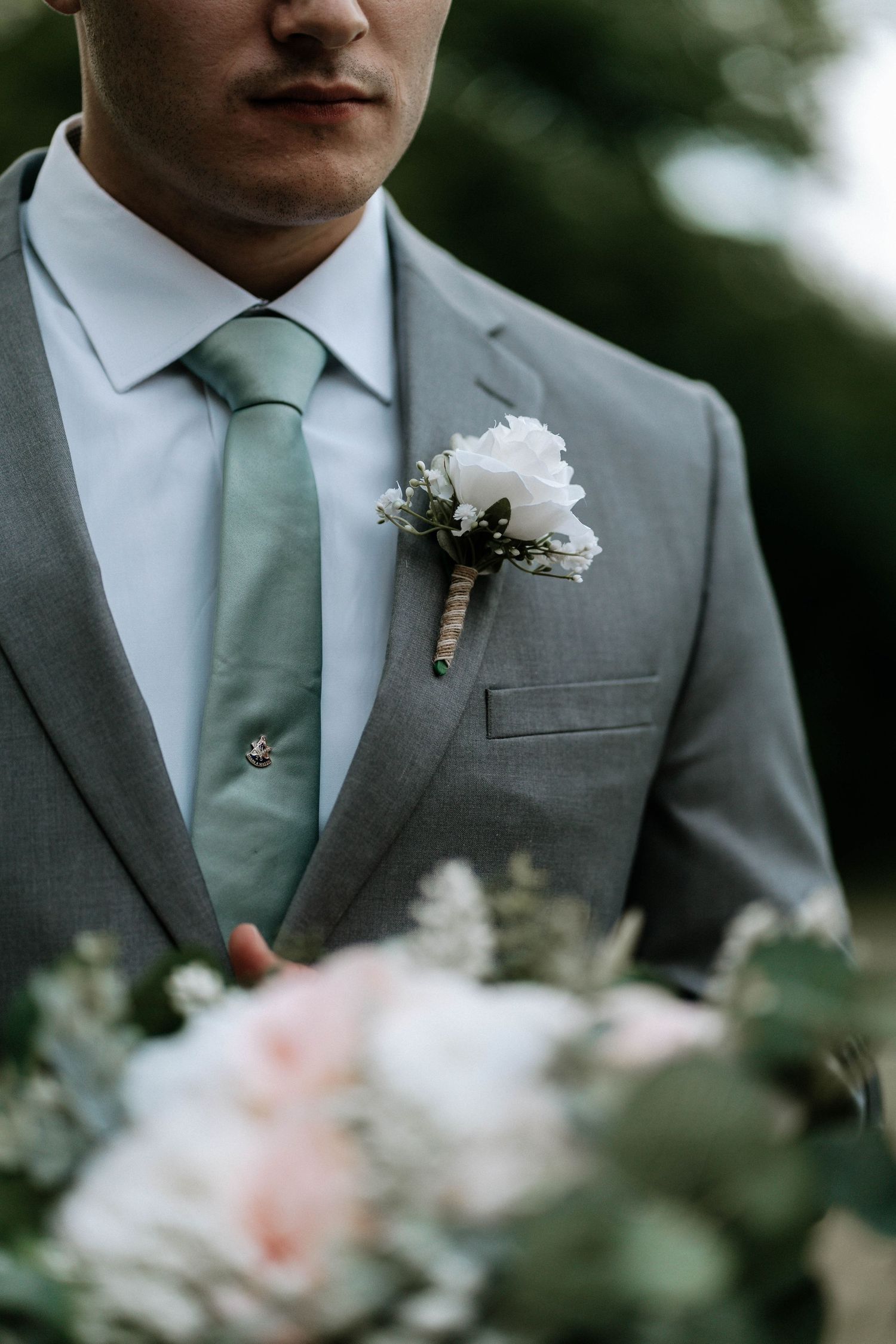 Groom in gray suit with sage green tie and white floral boutonniere holding a blush pink wedding bouquet.