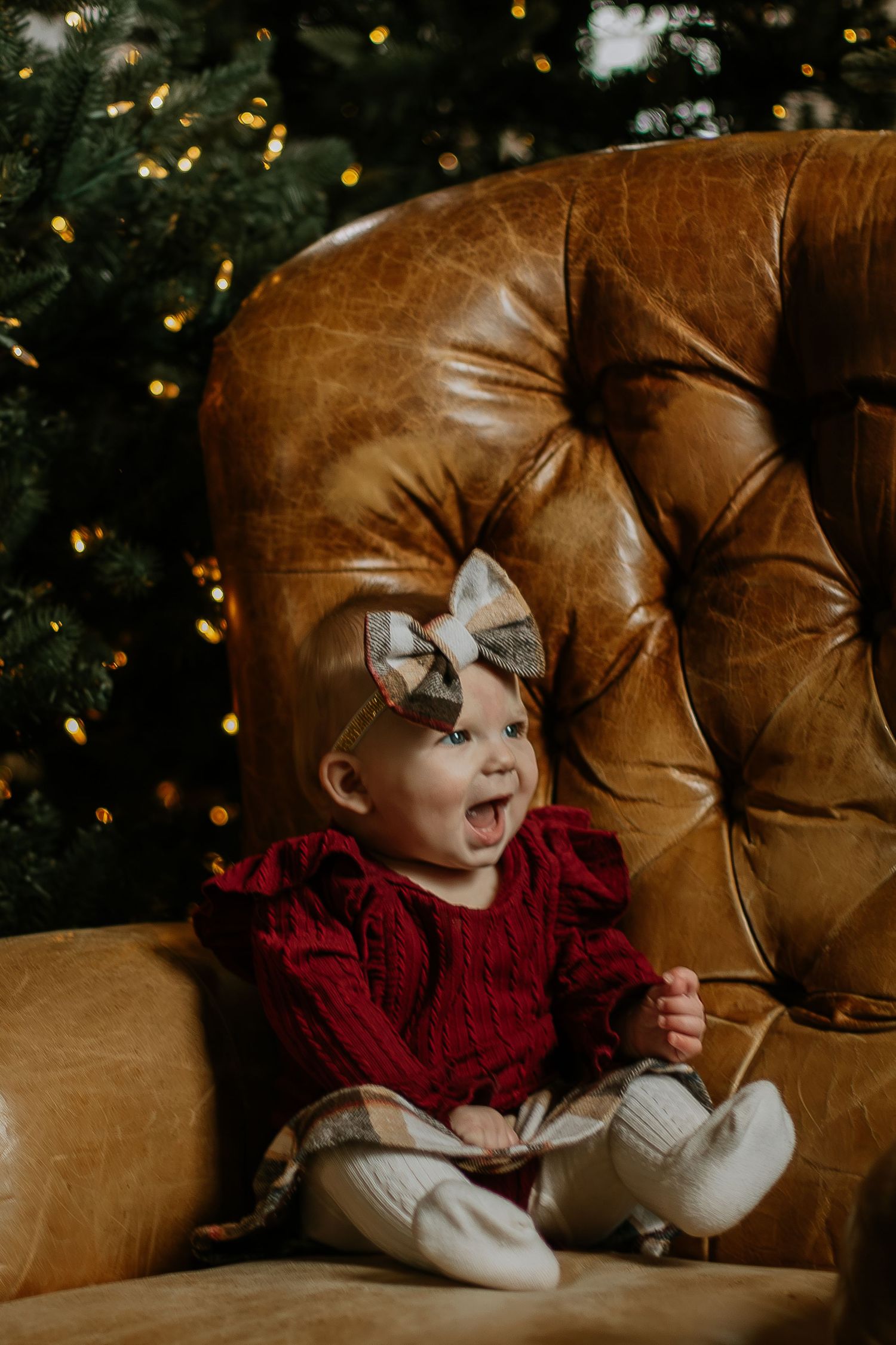 Happy baby girl in red dress sitting in leather chair near Christmas tree in Cincinnati, Ohio.