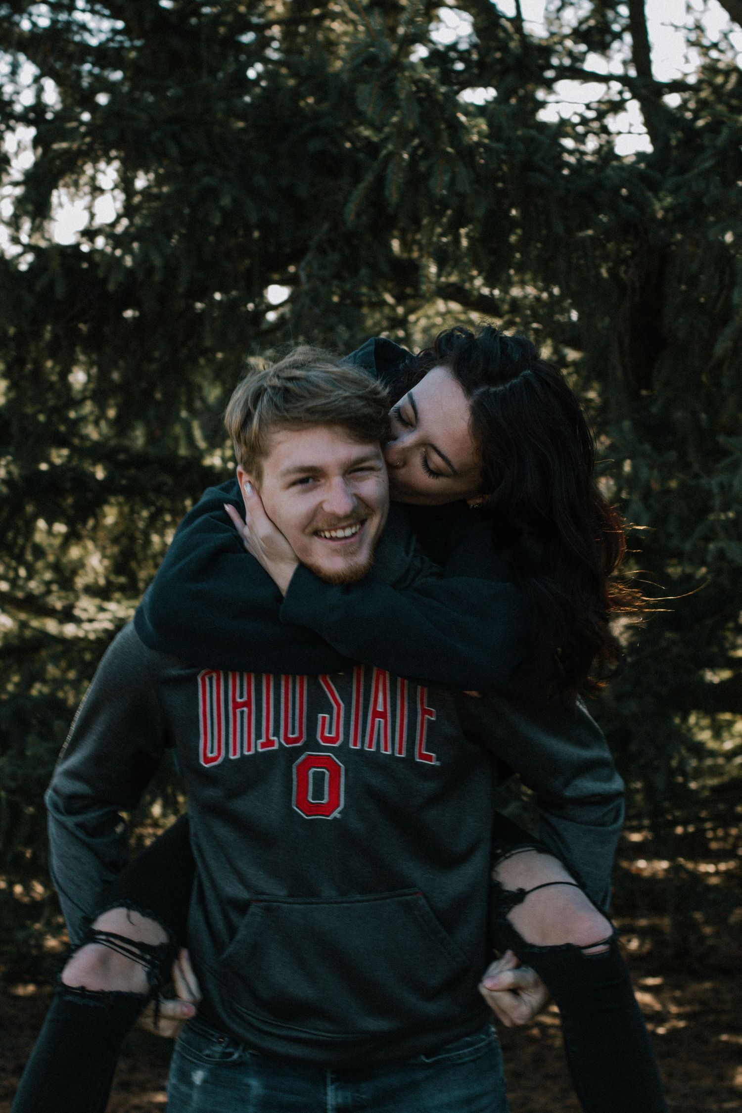 Woman kissing man on cheek from behind in piggyback pose, man wearing Ohio State hoodie outdoors in Dayton, Ohio